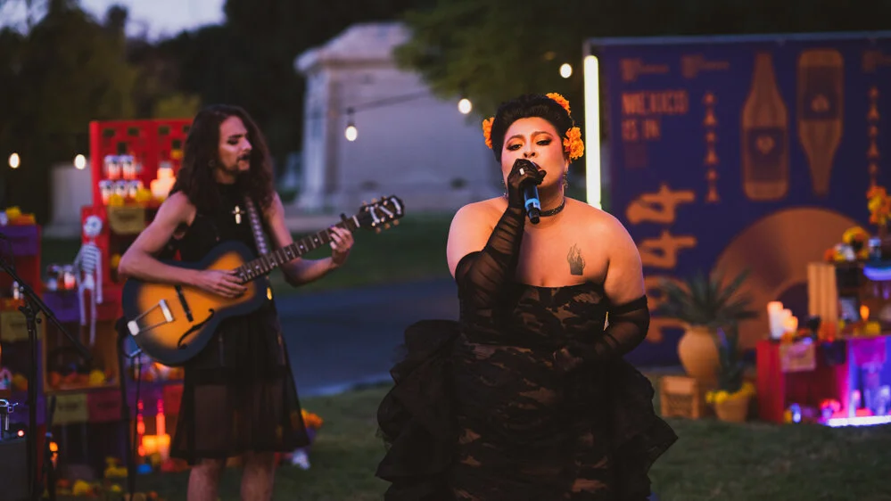 Mujer cantando con micrófono y vestida de negro, acompañada por un hombre tocando la guitarra, en un escenario decorado con flores y luces en un evento al aire libre por la tarde.