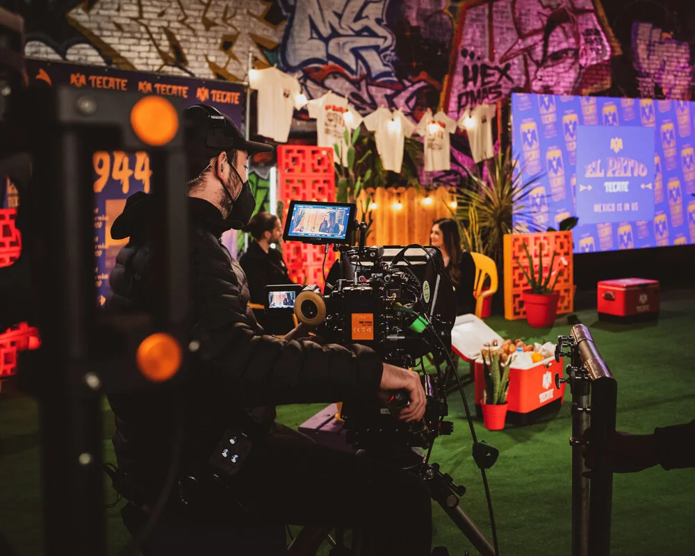 Camarógrafo grabando una entrevista en un set decorado con paredes de graffiti y plantas, con fondo azul y pantallas que muestran logotipos y texto en español.