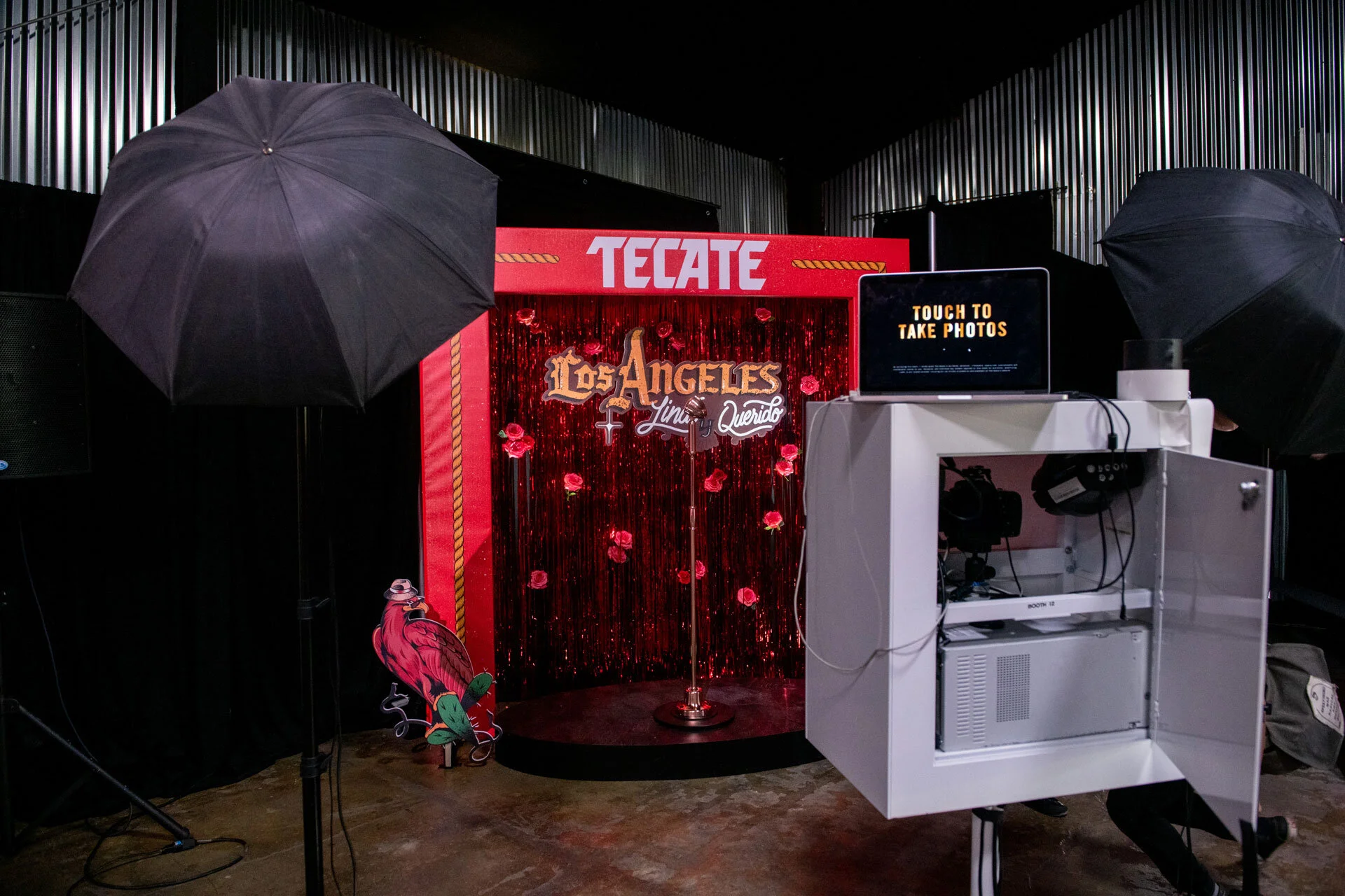 Estudio fotográfico con fondo decorativo rojo y luces, equipado con un fotomatón y un monitor que dice 'Touch to take photos'. Hay dos paraguas de estudio y un cartel con un loro en la parte inferior izquierda.