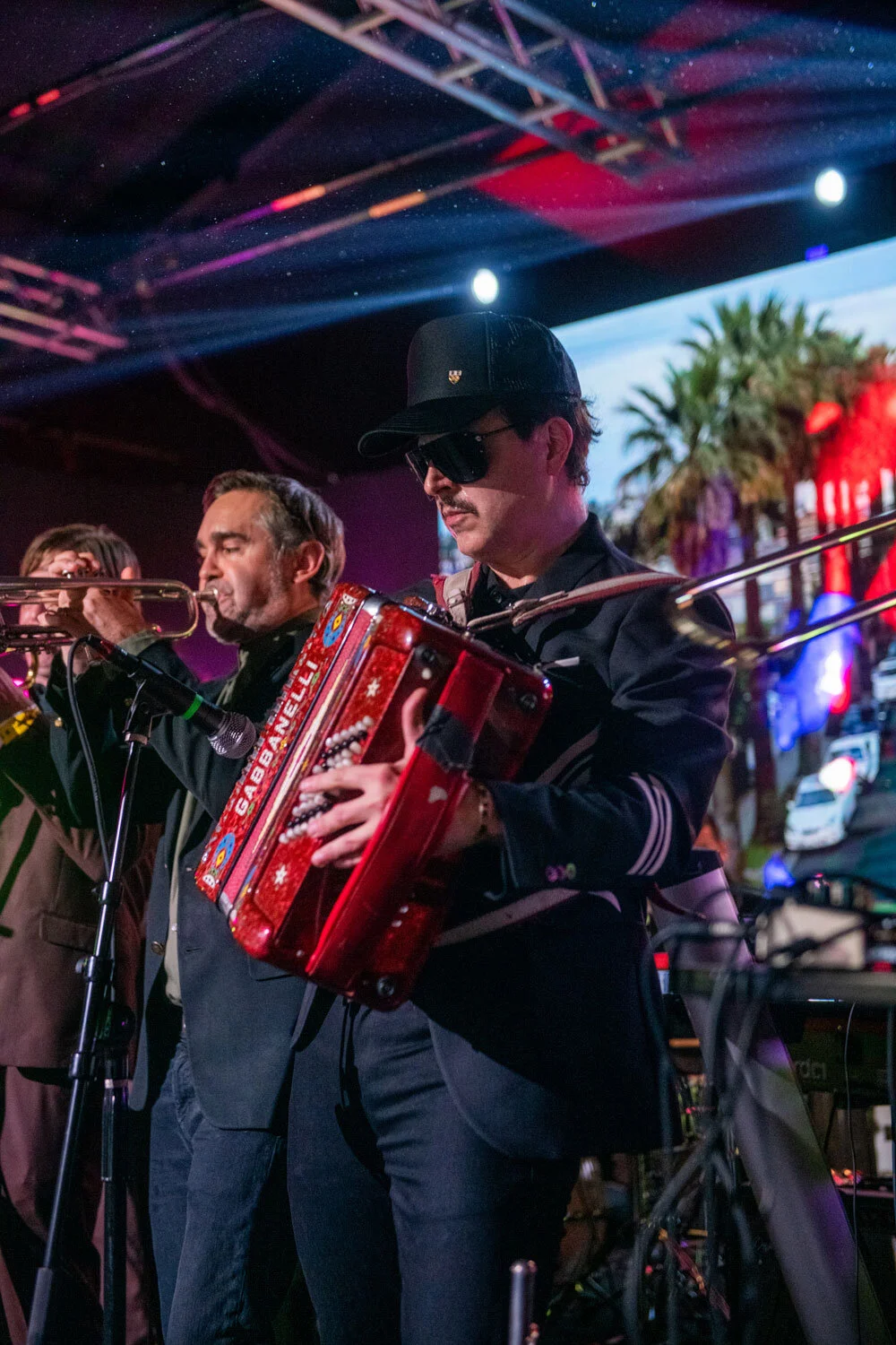 Músico tocando acordeón en concierto en la calle, con otros músicos y un fondo con palmeras y luces de neón.
