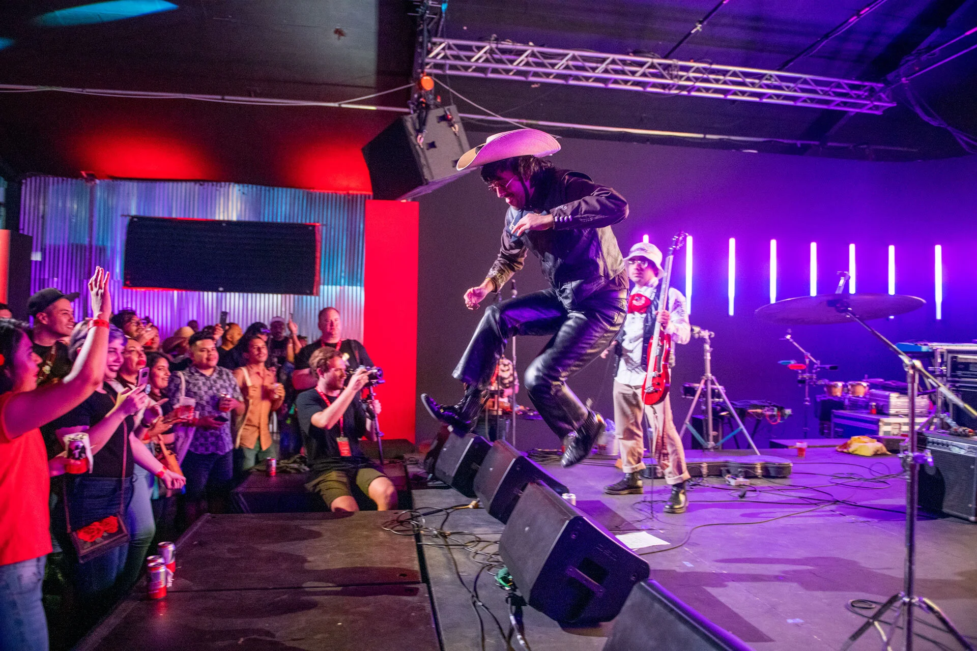 Un artista en un espectáculo en vivo saltando sobre el escenario con una banda tocando música, audiencia admirando y tomando fotos en un ambiente con luces de neón y equipo de sonido.