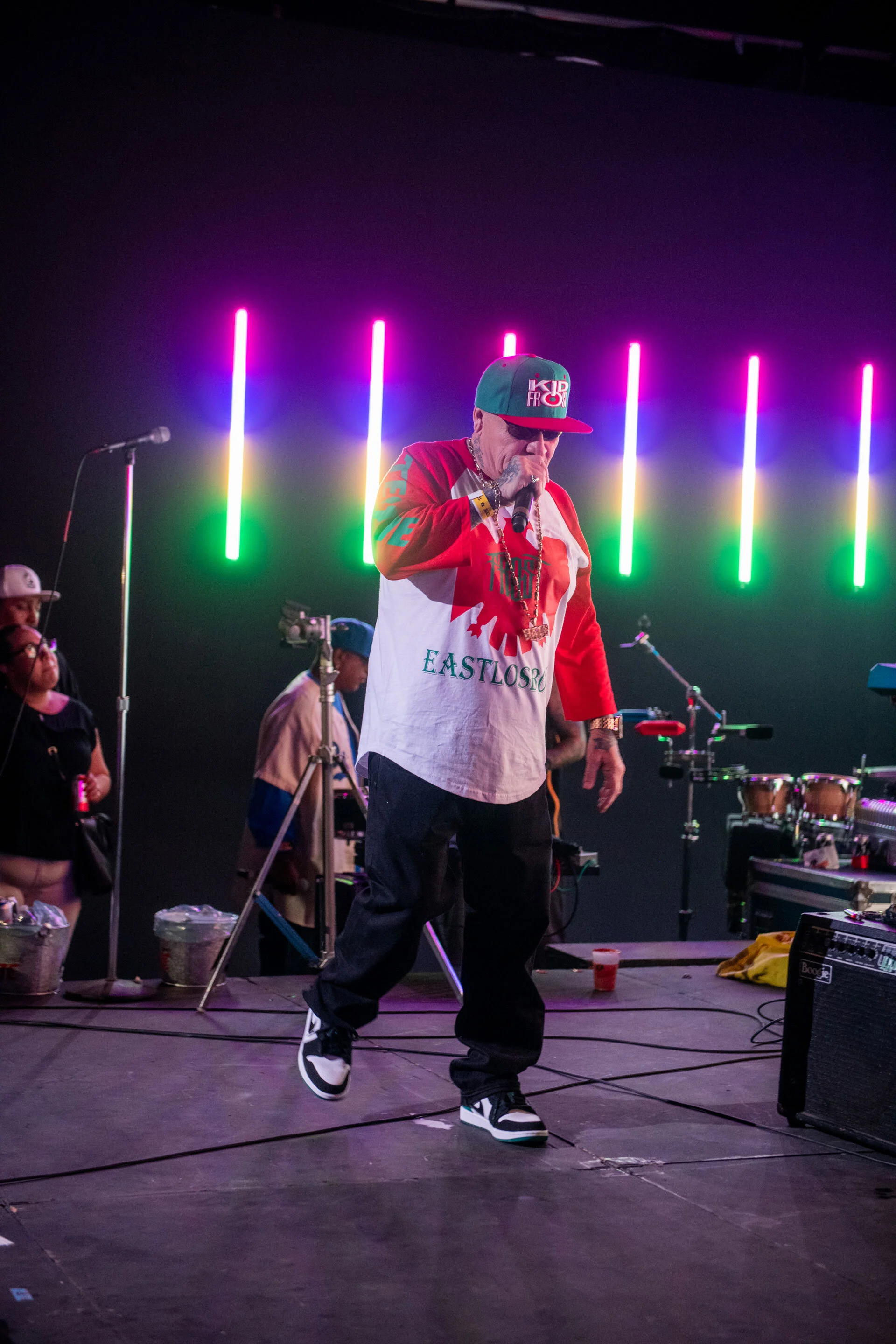Un hombre vestido con ropa colorida y gorra, cantando en un micrófono en un escenario con luces de neón de colores.