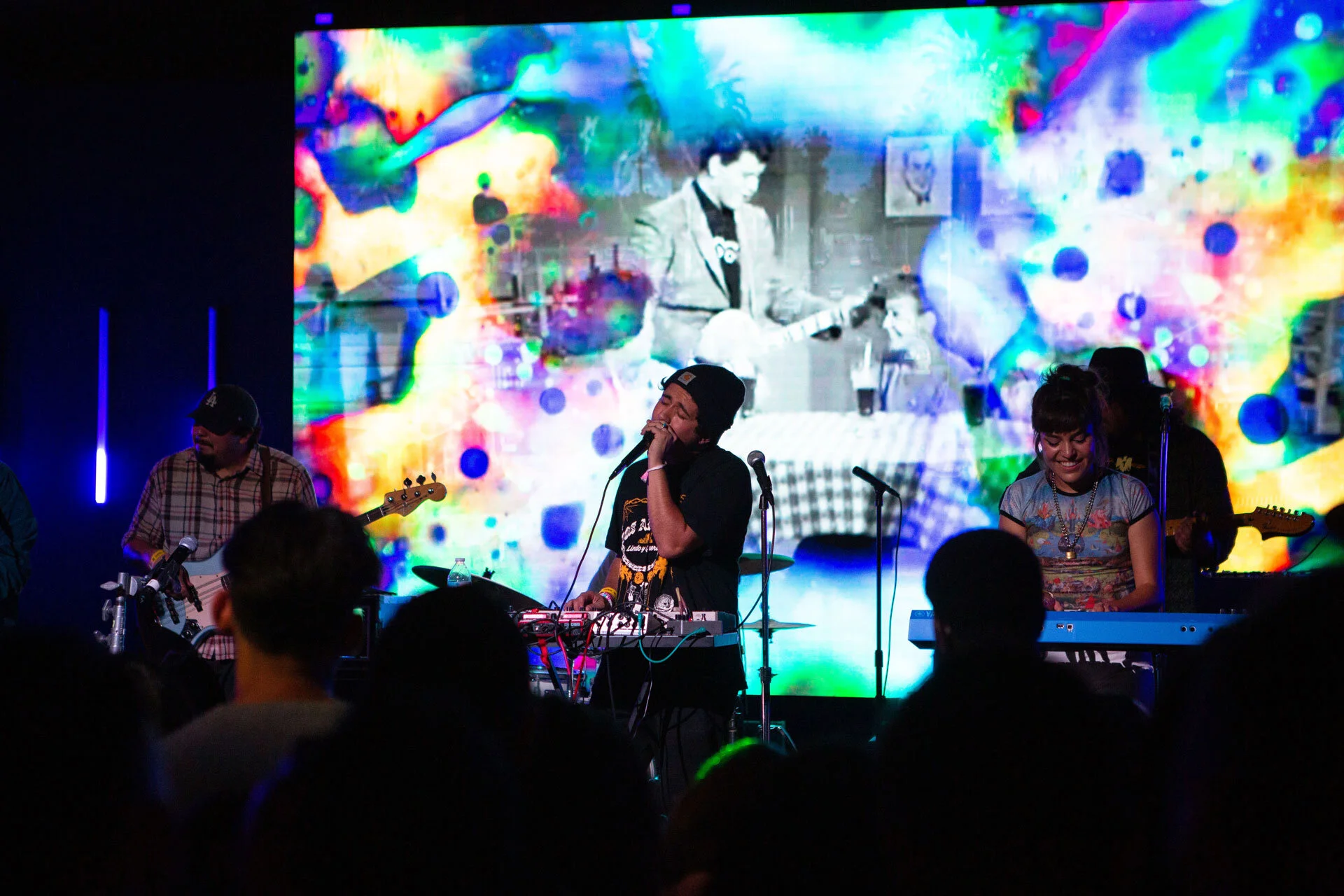 Conjunto de músicos en un escenario durante una presentación en vivo, con un fondo digital colorido y una audiencia en primer plano.