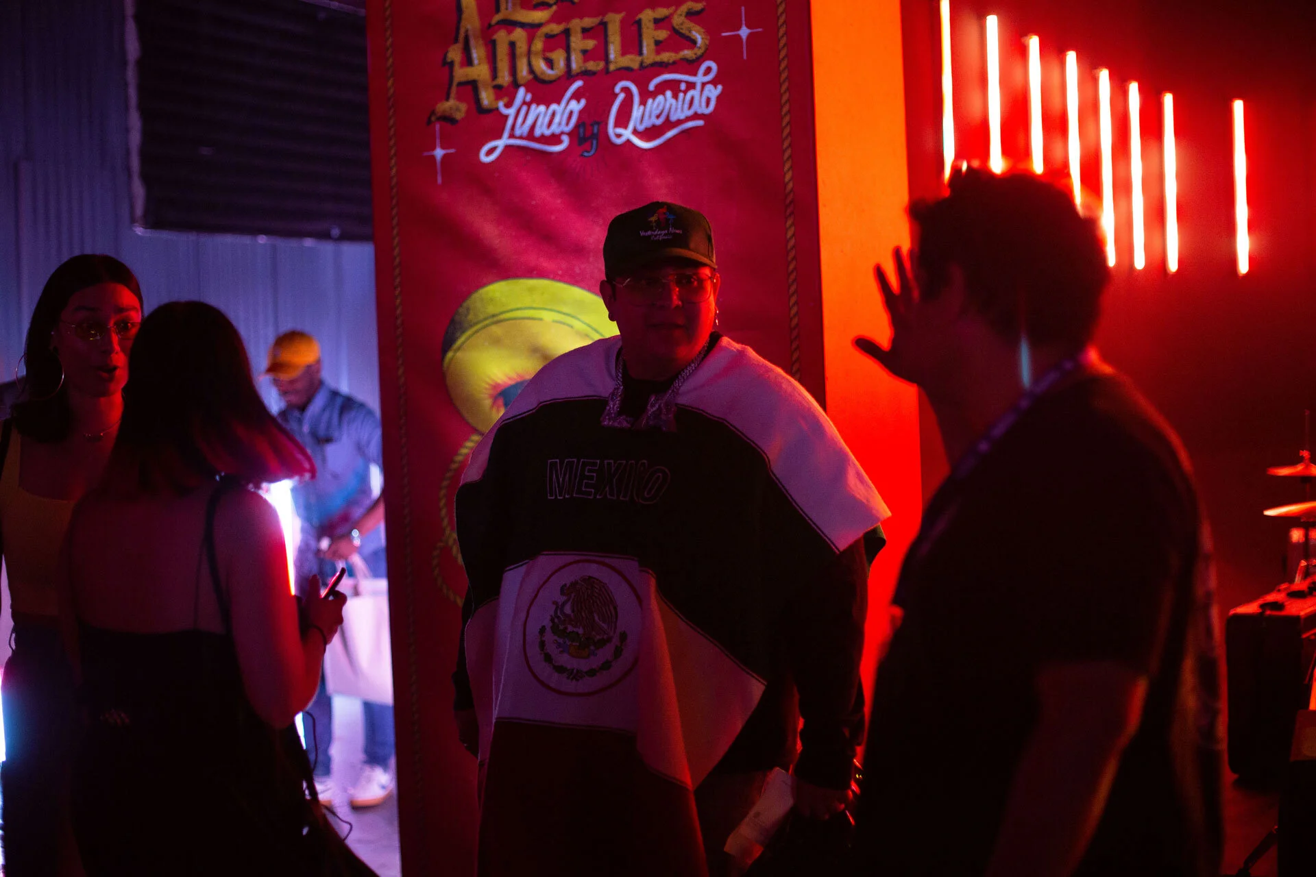 Personas en un evento con iluminación de neón rojo, charlando cerca de un cartel que dice 'Ángeles Linda y Querido', con una de ellas vistiendo una chaqueta de México y la bandera mexicana.