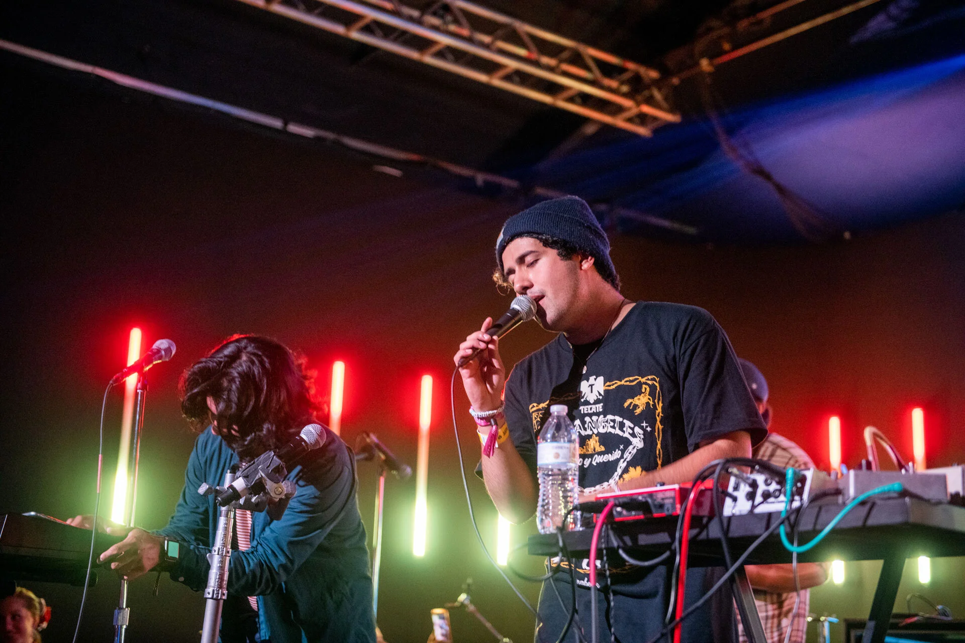 Dos músicos en un escenario: uno con una camiseta negra y una gorra negra, tocando con un micrófono, y otro con un teclado y una prótesis en un brazo, en un fondo con luces de neón rojas y verdes.
