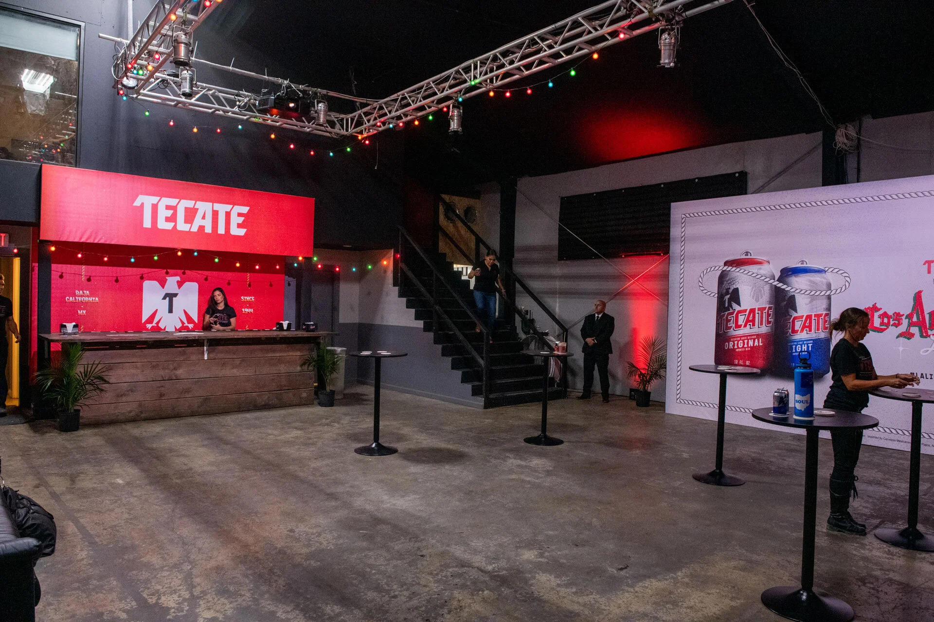 Interior de un establecimiento con área de recepción decorada con un cartel grande que dice Tecate, con mesa de recepción de madera, plantas en las esquinas, y varias mesas pequeñas con botellas de agua y vasos. Hay personas, una tras la recepción y 