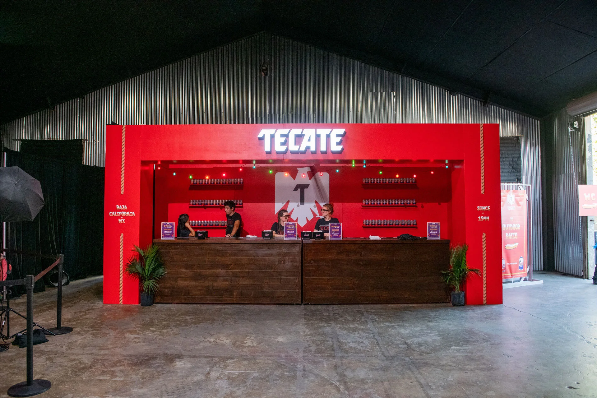 Mostrador de un puesto de comida con fondo rojo y logo de Tecate, dos mujeres y un hombre atendiendo, decoraciones de plantas y luces de colores.