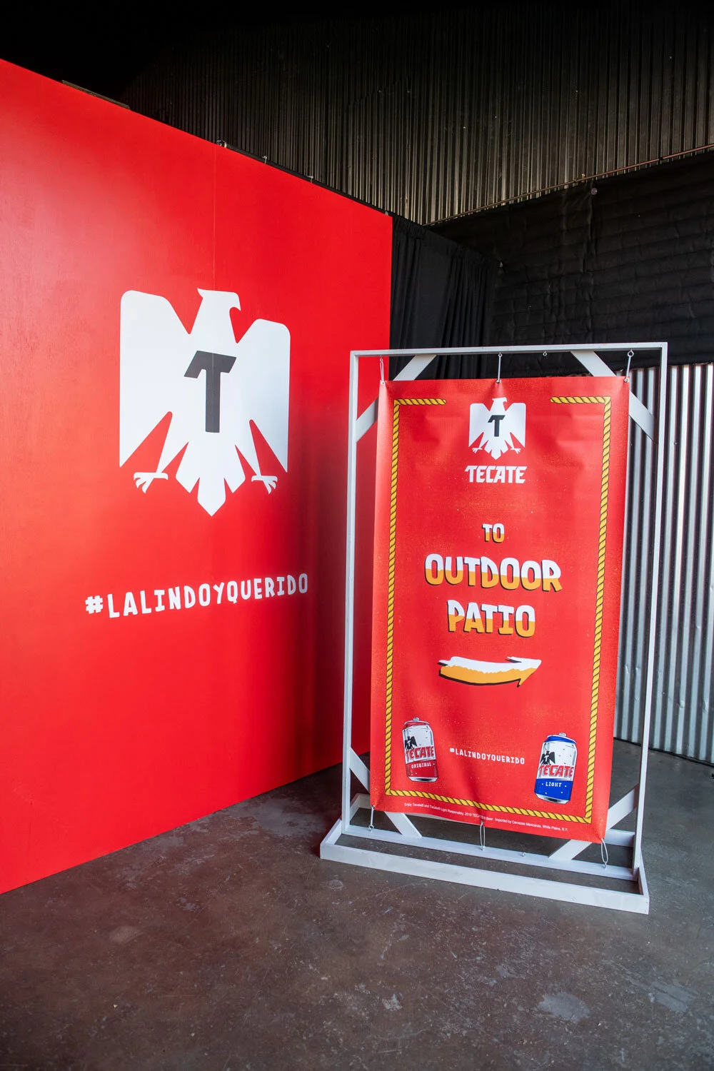 Cartel rojo con logo de águila y texto en inglés y español que dice 'a TECATE to outdoor patio' y '#LALINDOYQUERIDO', con ilustraciones de latas de cerveza.