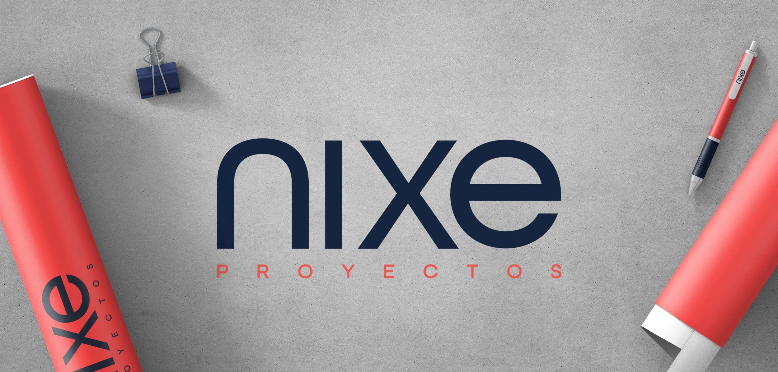 Logo de 'Nixe Proyectos' en medio de una mesa gris rodeado de papelería, incluindo dos rollos de papel, un bolígrafo y una pinza metálica con clip azul.
