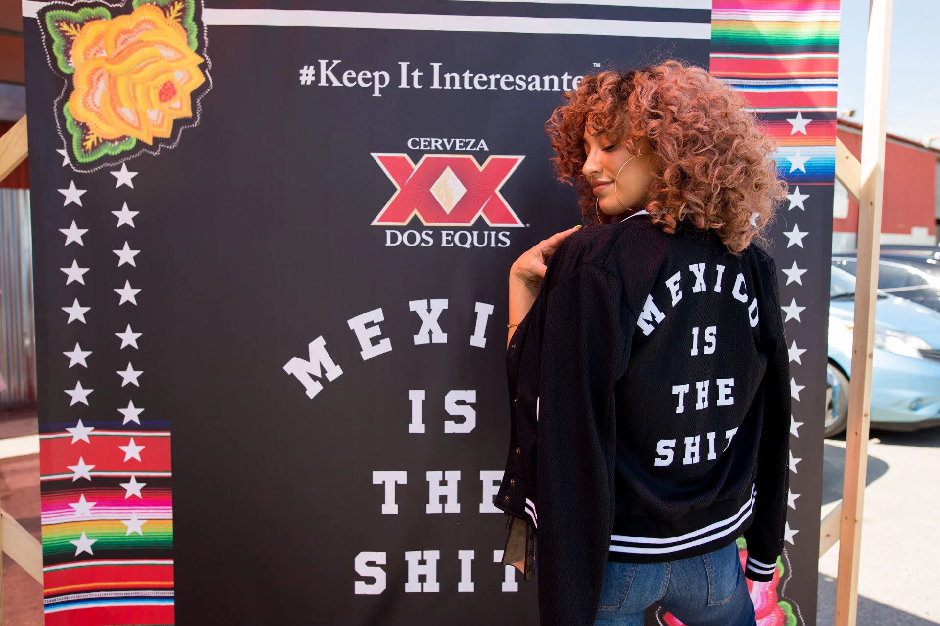 Mujer de cabello rizado pelirrojo posando frente a cartel con texto en inglés, en un evento al aire libre.