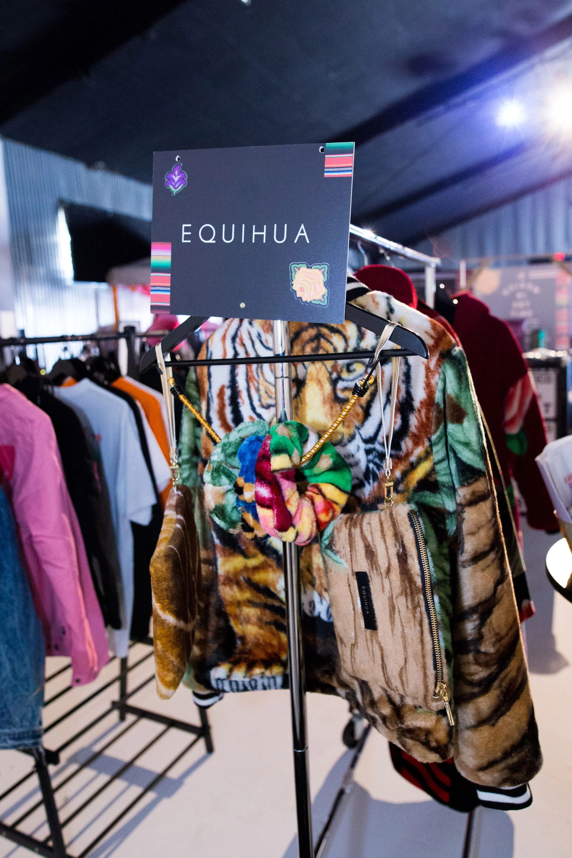 Ropa y accesorios de la marca Equihua en una tienda, incluyendo un abrigo con estampado de tigre, un lazo de tela colorido, y otros artículos en un perchero bajo una señal con el nombre de la marca.
