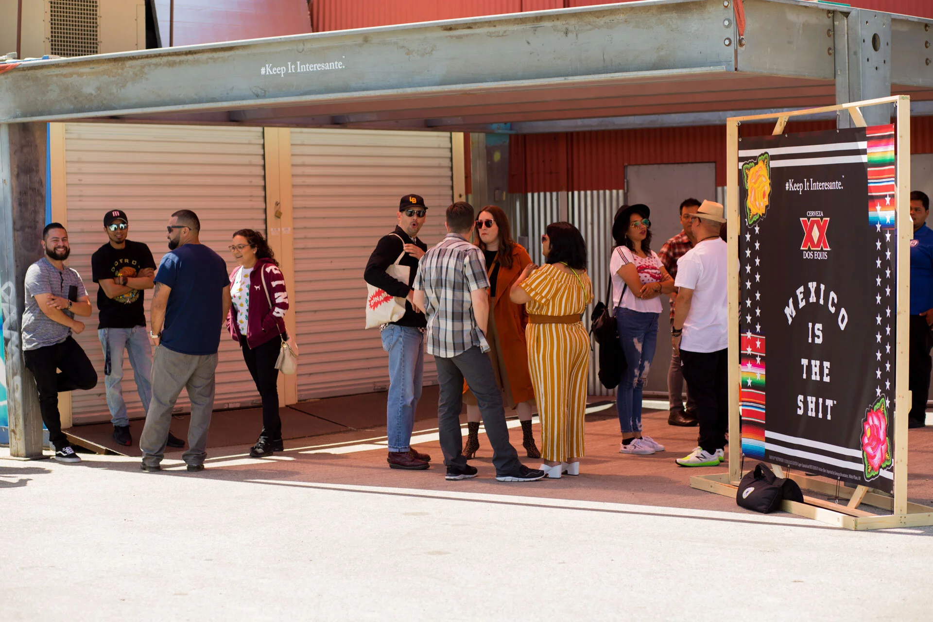 Grupo de personas haciendo fila en un lugar público con cartel que dice "México as the shit" en inglés y otra cartel en la esquina que dice "Keep it interesante".