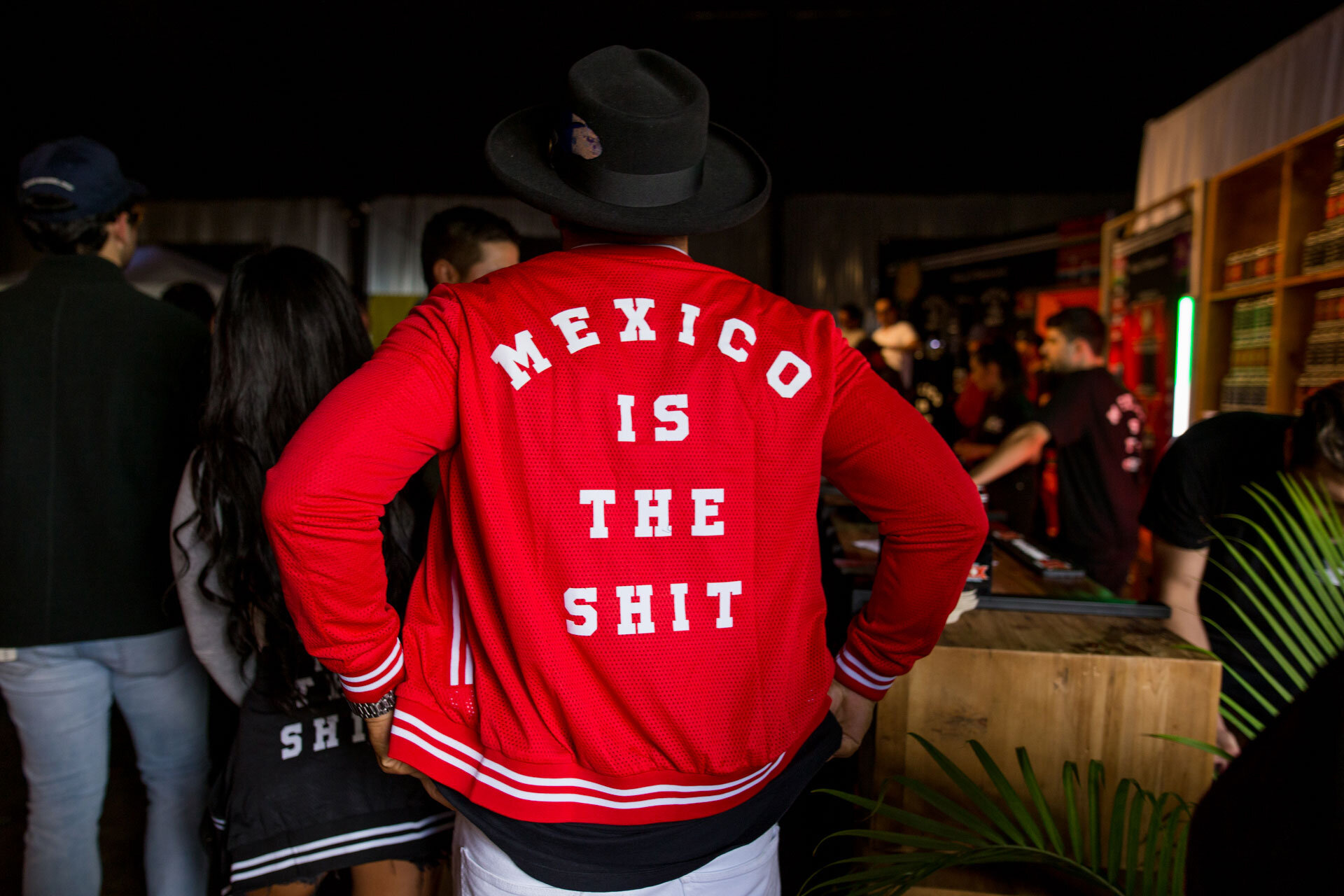 Persona con chaqueta roja que dice 'México is the shit' en una fiesta o evento, con gente y estanterías de libros al fondo.