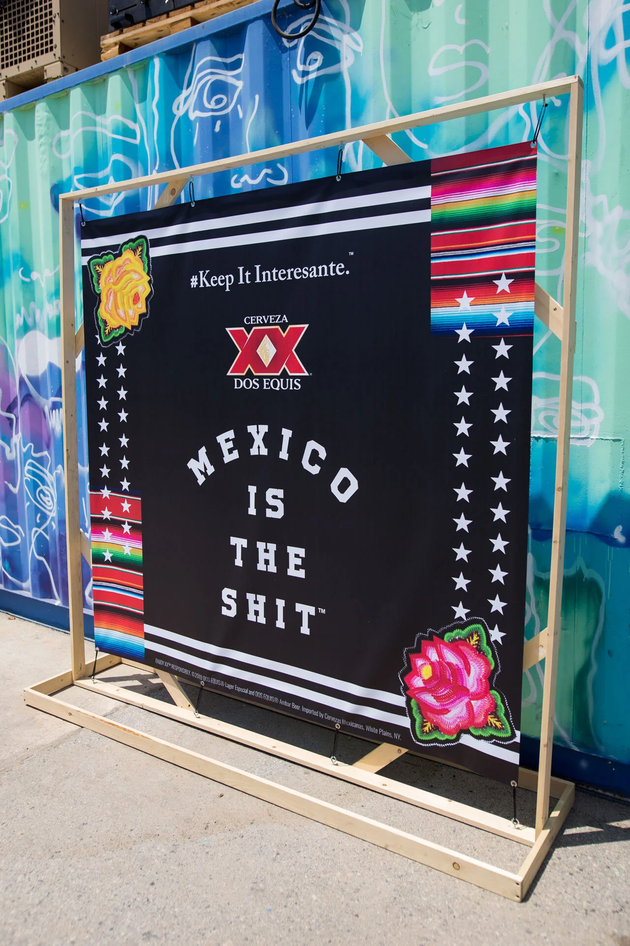 Cartel publicitario en español con fondo negro que dice "México is the shit" y tiene logos de cerveza, rosa y decoraciones coloridas, ubicado en un marco de madera en la calle con un graffiti de fondo.