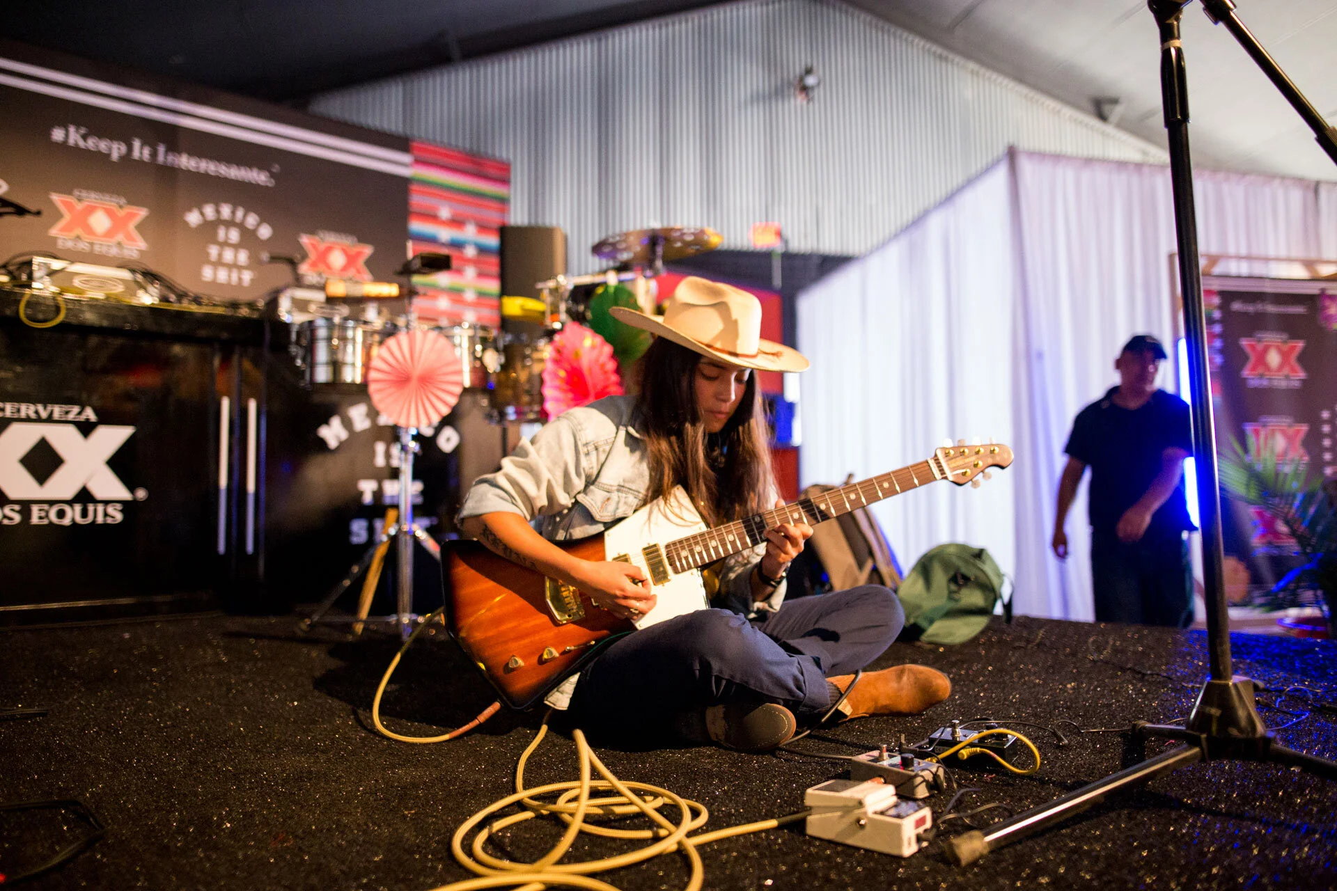 Una mujer tocando la guitarra en un escenario, usando sombrero y ropa casual, con equipo musical y decoraciones coloridas alrededor.
