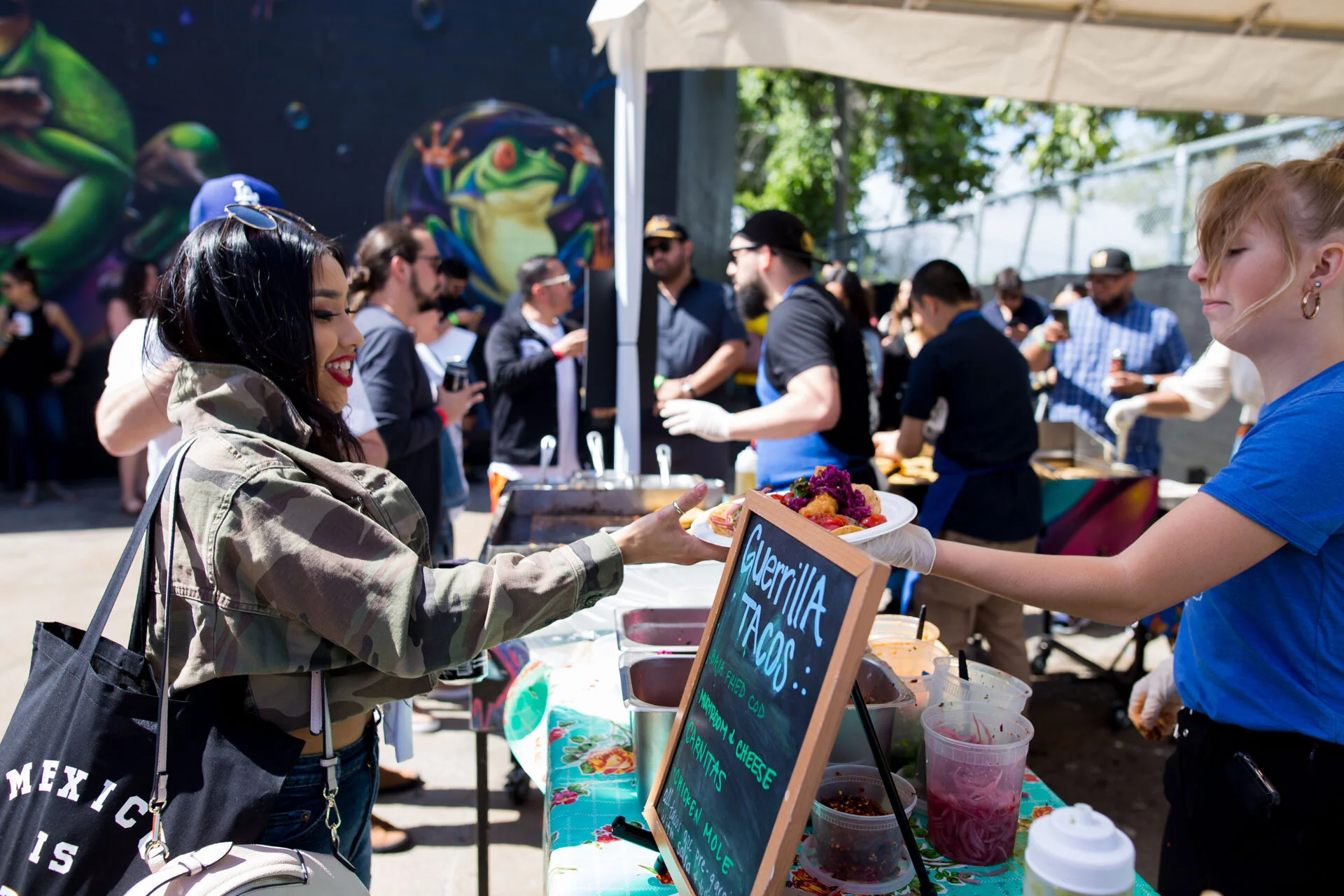 Mujer con chaqueta de camuflaje recibe un plato de comida en un puesto de comida mexicana en un evento al aire libre con otras personas en el fondo, paredes con arte de ranas y varios recipientes con ingredientes.