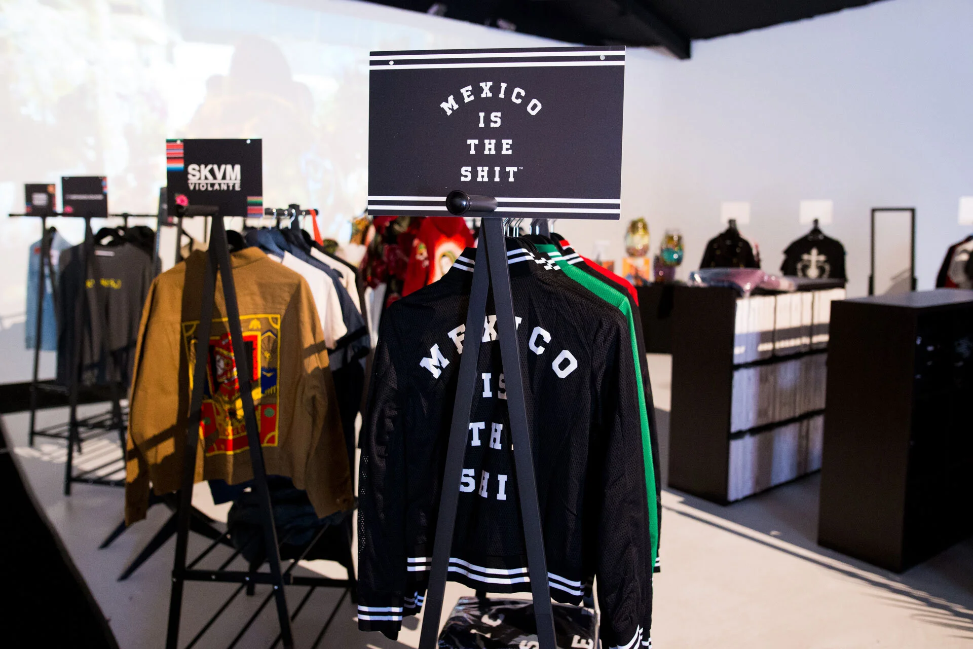 Ropa en una tienda. Hay chaquetas de diferentes colores, incluyendo una negra con letras blancas que dice 'Mexico is the shit'. Un cartel en la parte superior de las chaquetas también dice 'Mexico is the shit'.