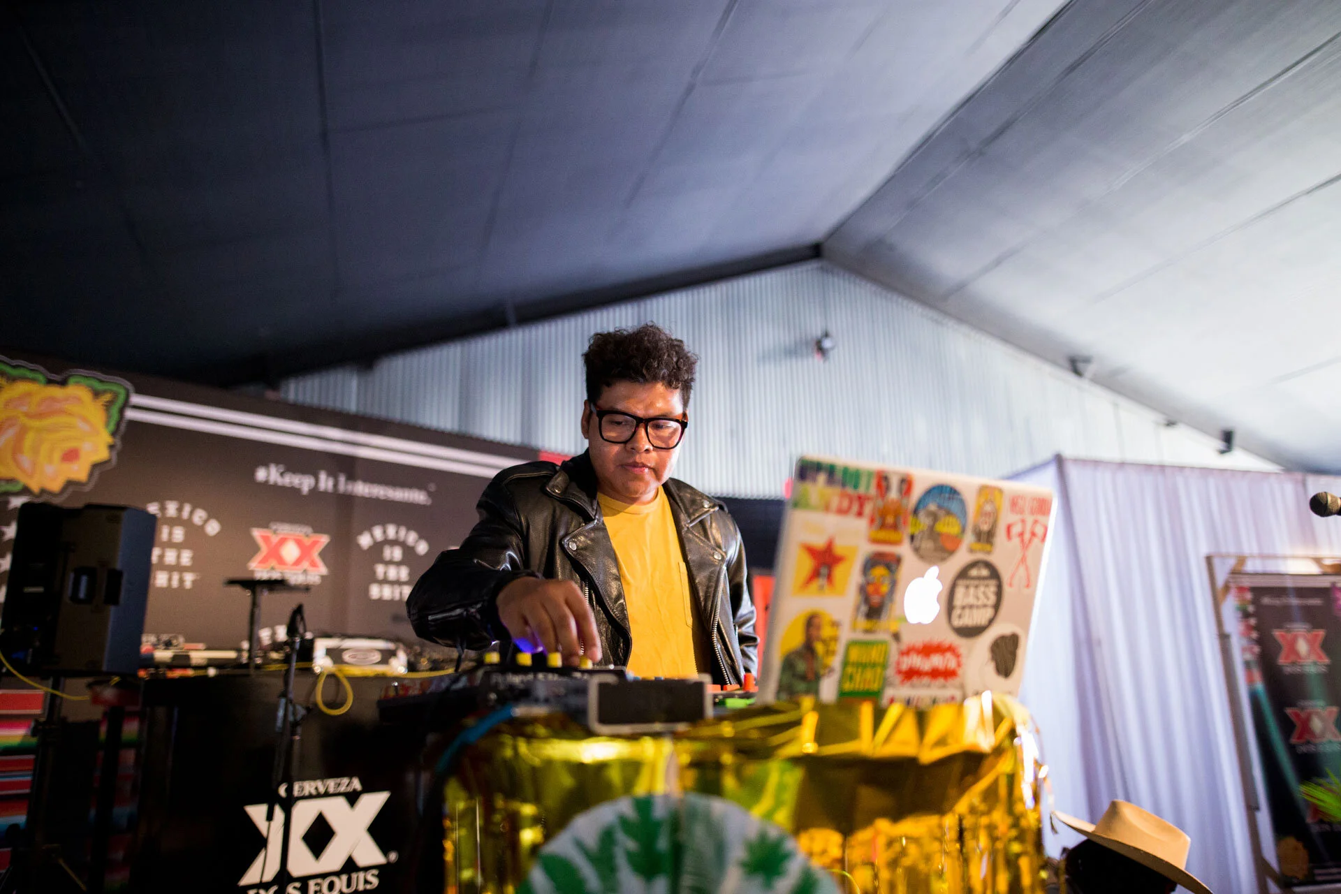 DJ mezclando música en un evento con decoraciones coloridas y un cartel de patrocinador en el fondo.