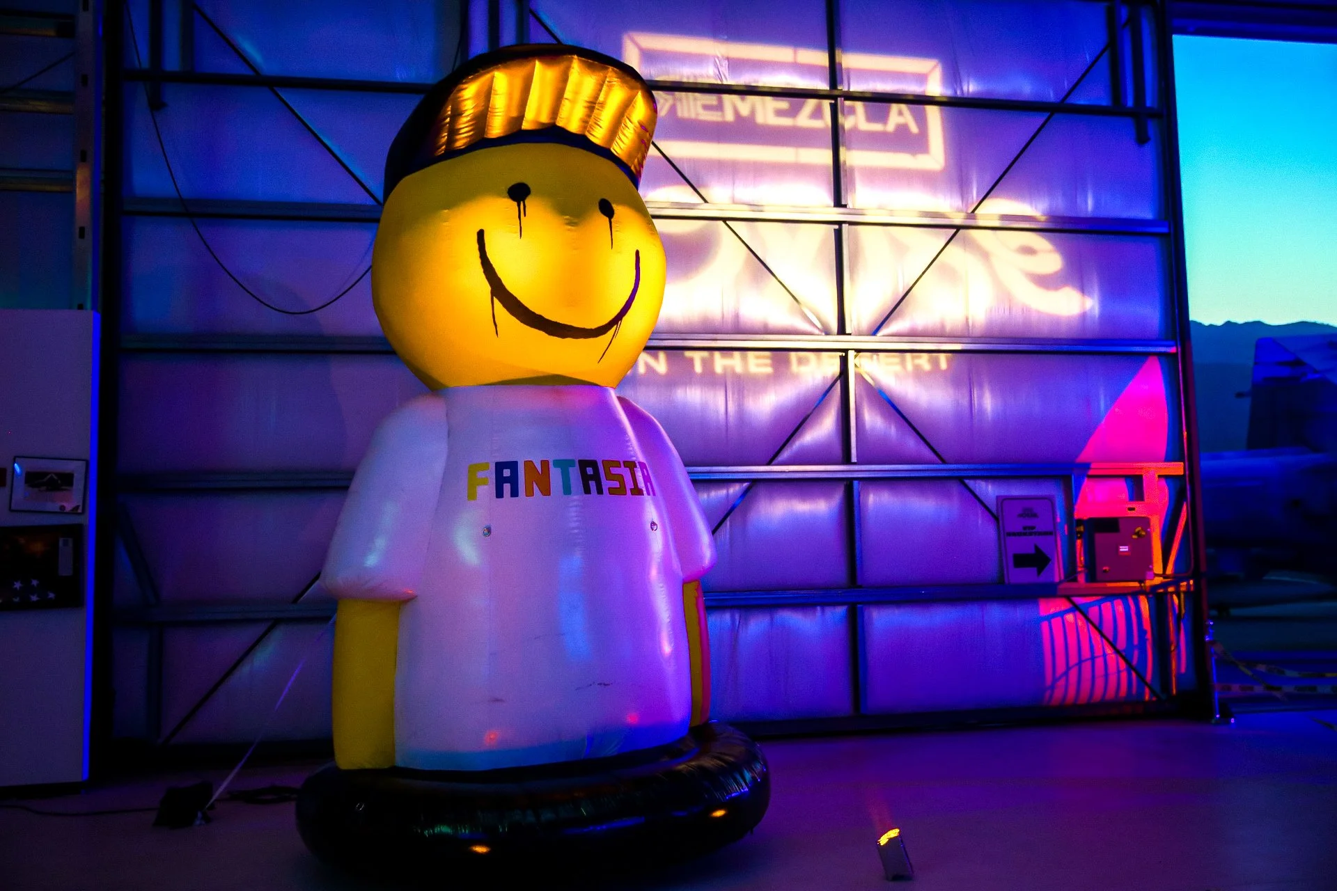 Una figura inflable con cabeza amarilla y sonrisa, con un gorro dorado, vestida con camiseta blanca que dice 'FANTASIA', en un espacio con luces de colores y un fondo metálico, en lo que parece ser un evento nocturno.