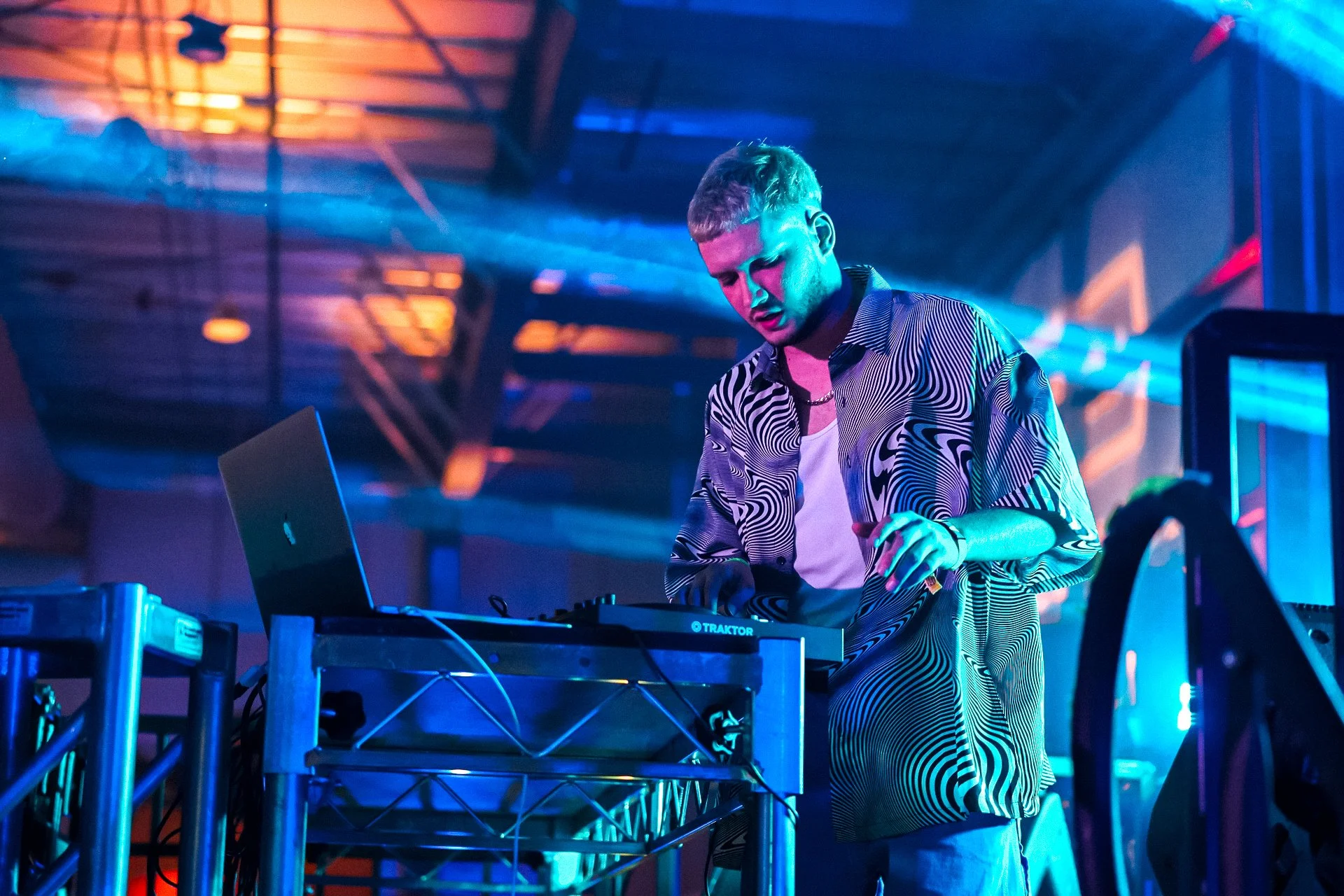 Un DJ con camiseta de patrones, tocando en una cabina con equipo de mezclas y una laptop en un ambiente con luces azules y naranjas durante un evento o fiesta.