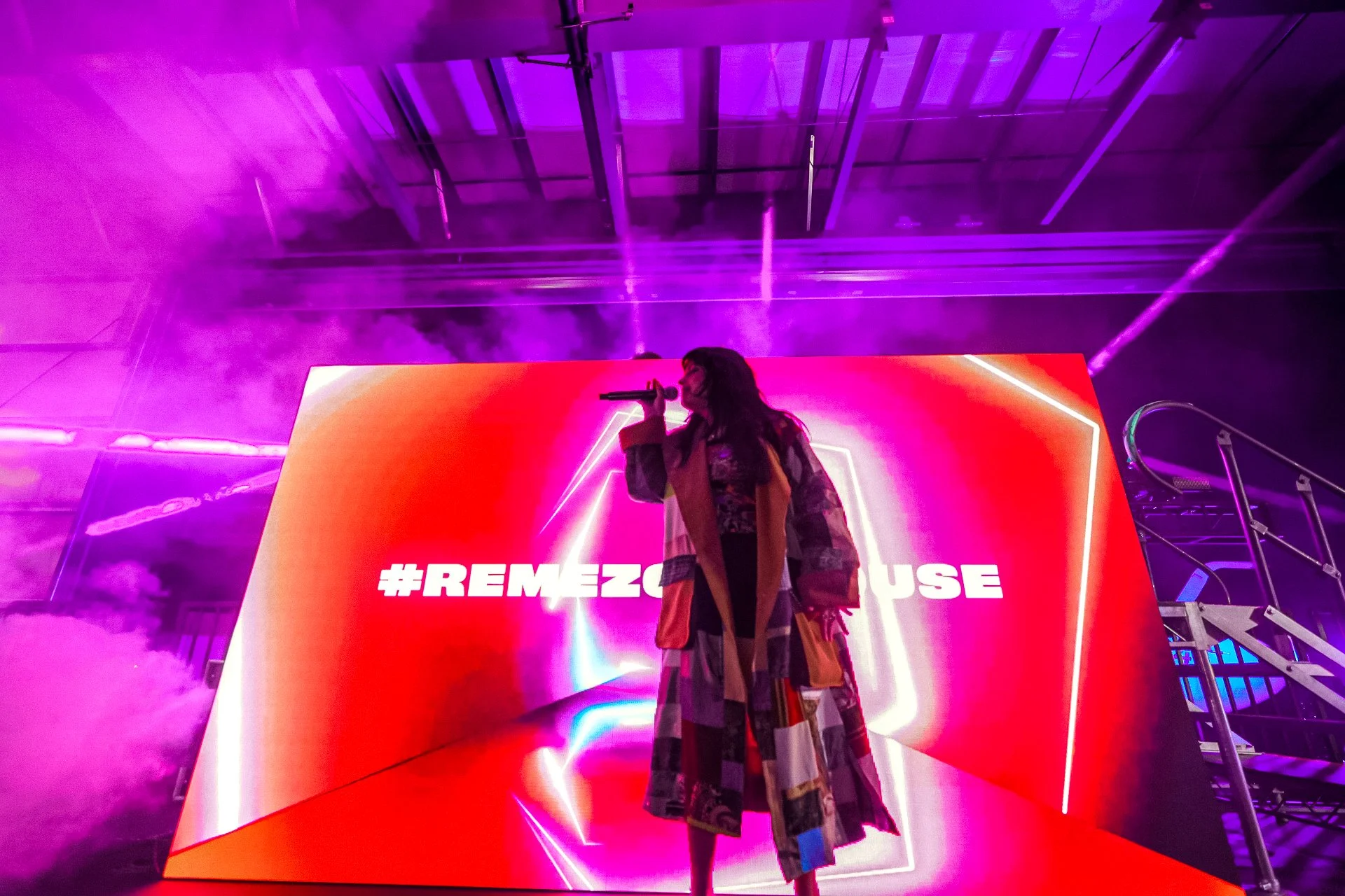 Una mujer con un abrigo colorido sostiene un micrófono frente a una pantalla grande con el hashtag #REMEZO en un escenario iluminado con luces moradas y neón.