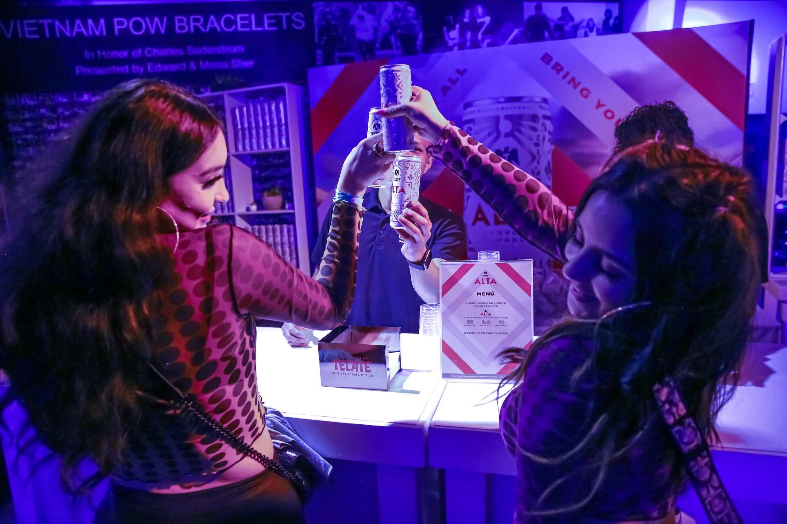 Tres mujeres celebrando con brindis en un bar, con iluminación azul y cartel que promociona pulseras de Vietnam POW.