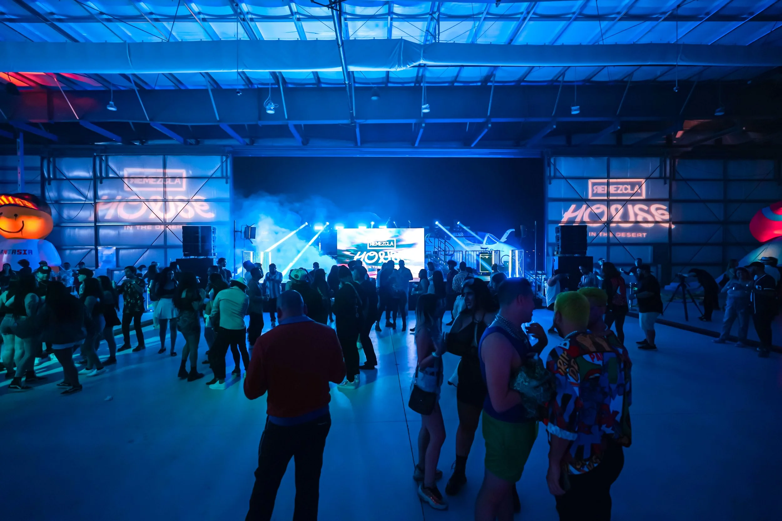 Escena en un evento musical con gente bailando y un escenario con pantallas y luces en azul, decorado con figuras inflables y letras que dicen "ReeZcl" y "HOU5".