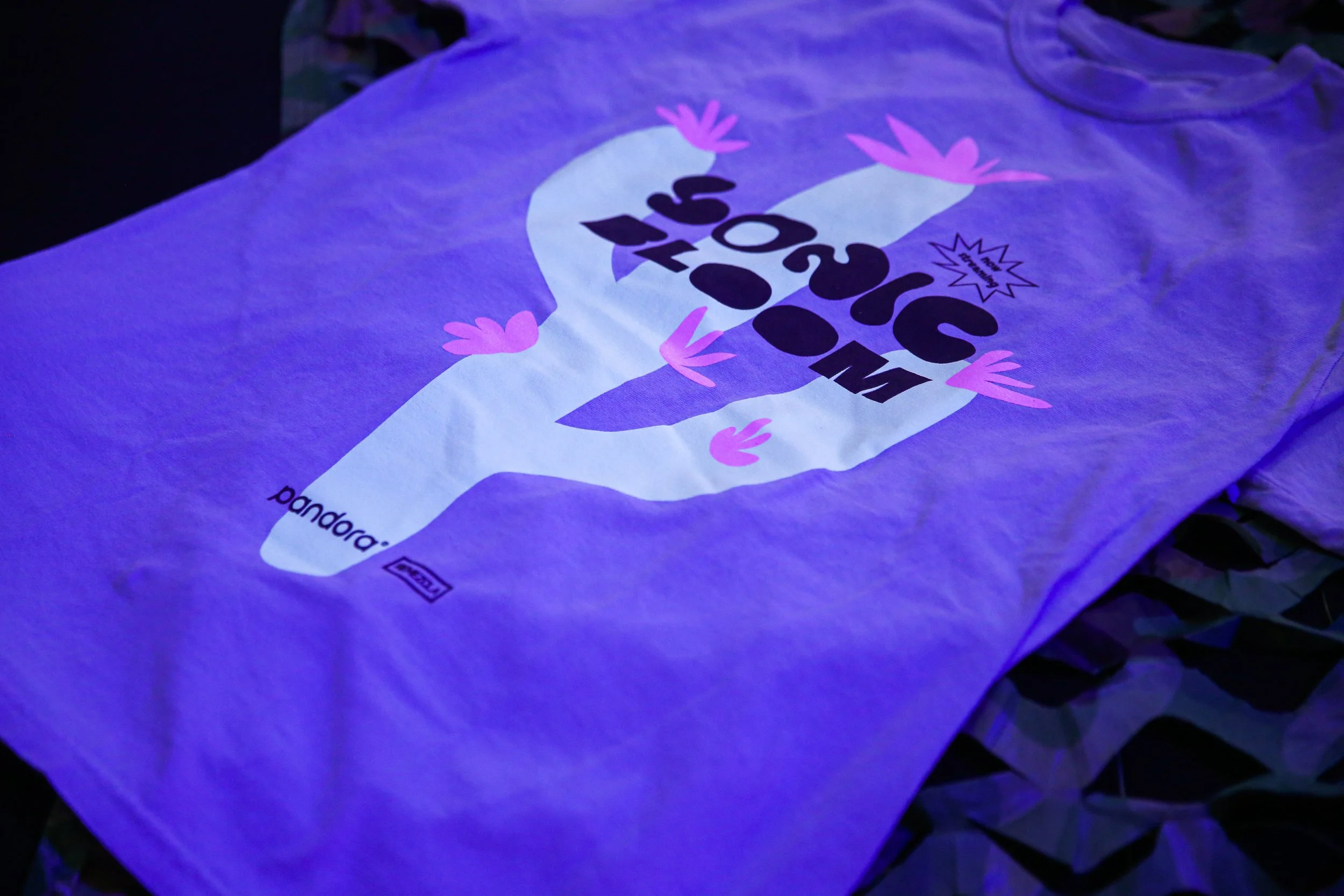 Camiseta blanca con texto y gráficos en color púrpura y rosa, que parece estar bajo luz ultravioleta, en un entorno oscuro.