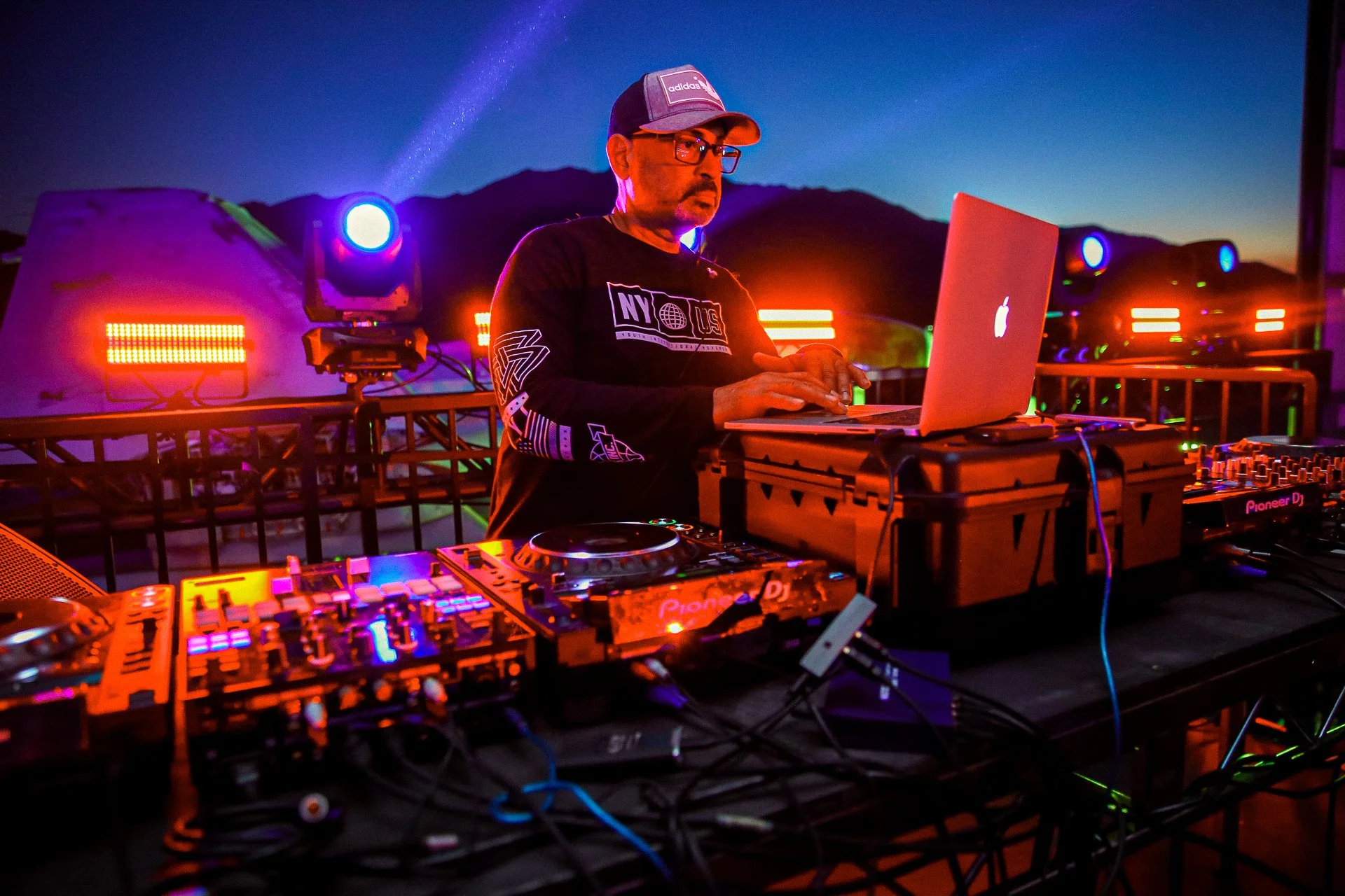 DJ en una cabina con equipo de música, luces de colores y un fondo de montañas durante la noche.