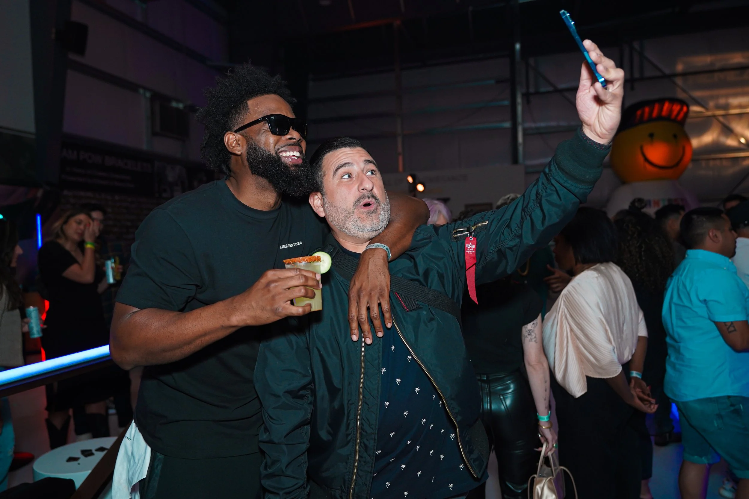 Dos hombres en una fiesta social toman una selfie, uno con gafas de sol y sosteniendo una bebida, el otro con expresión sorprendida, rodeados de otras personas en un ambiente animado.
