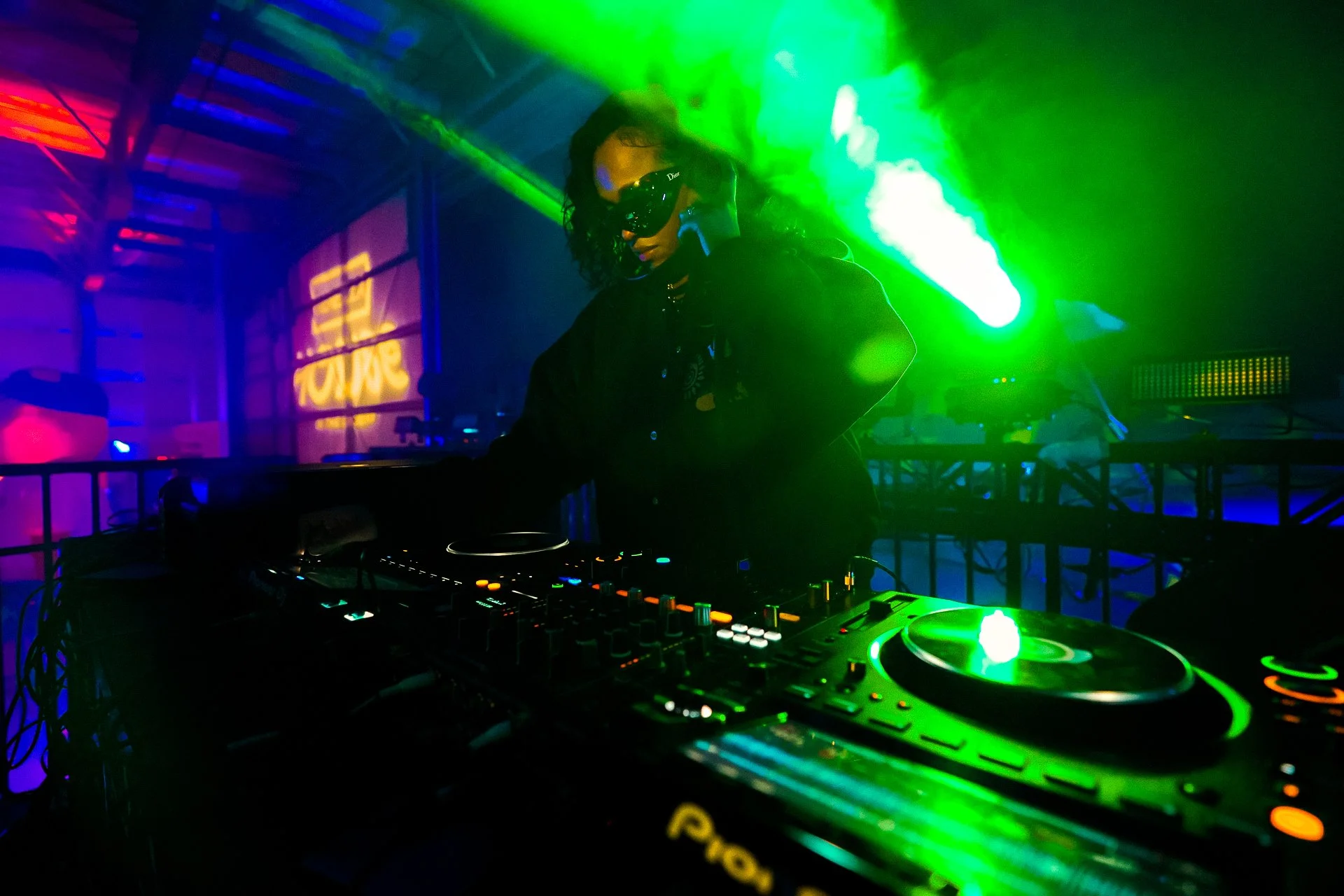 DJ en un club nocturno, con iluminación de colores verdes, azules y rojos, usando gafas de protección y operando equipo de sonido y mezcladoras.