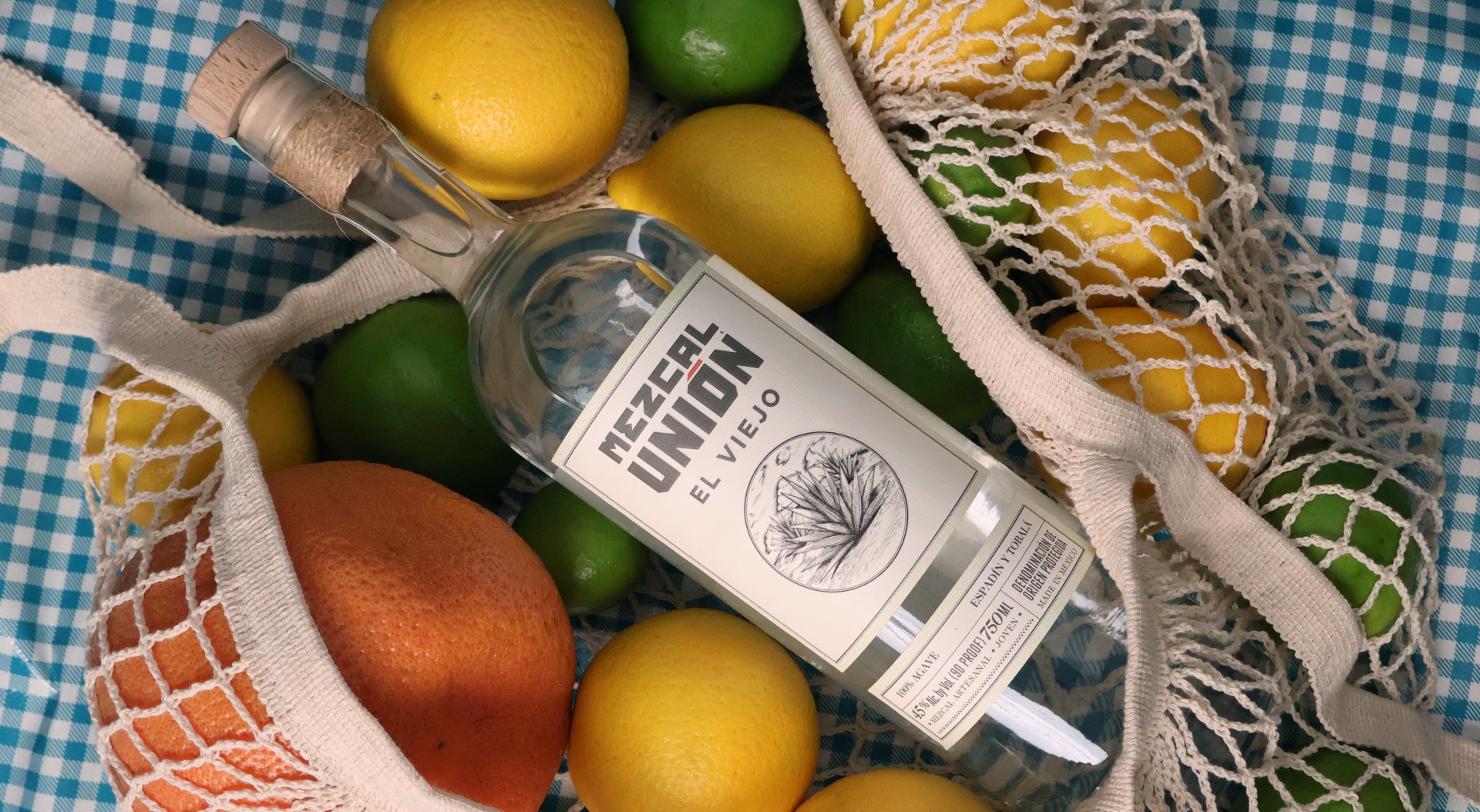Botella de tequila 'Mezcal Unidad', rodeada de frutas cítricas como limón, lima, naranja y un pomelo, en una malla de tela blanca sobre un mantel de cuadros azules y blancos.
