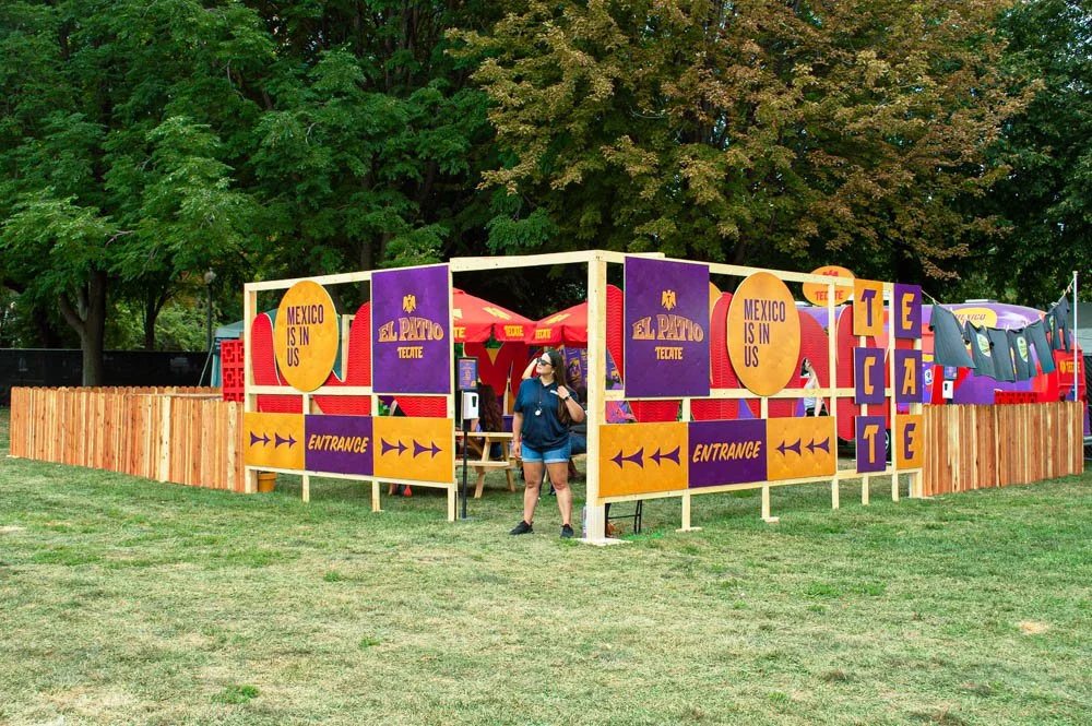 Stand decorativo con señalización en colores vibrantes que dice 'El Pato Tequila' y 'Mexico is in us', rodeado de árboles y con una mujer caminando cerca.