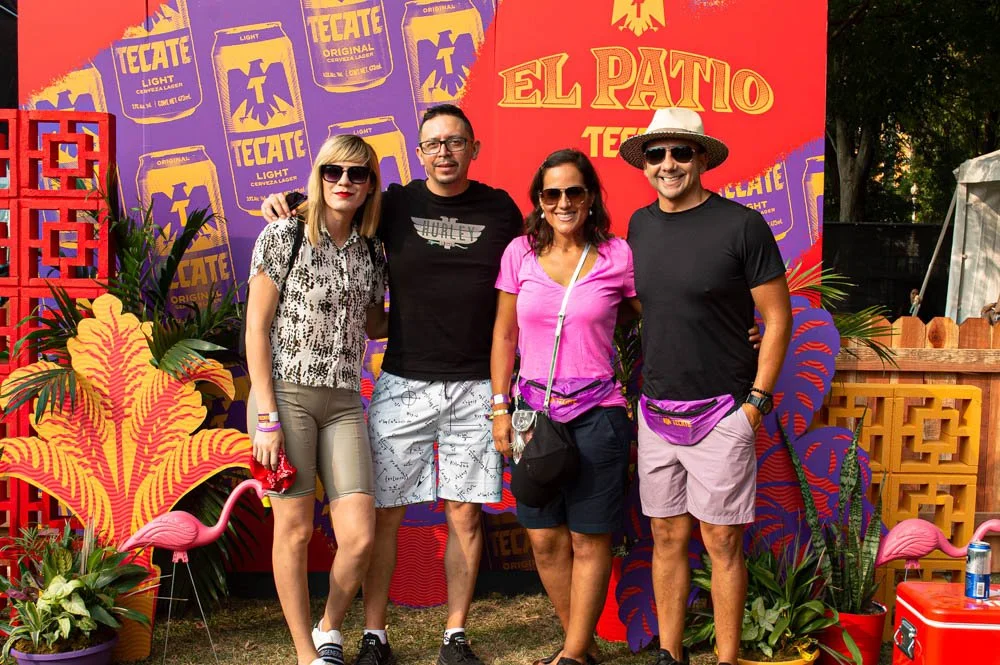 Cuatro personas de aspecto relajado posando en un evento con decoración tropical y banners de Tecate. Hay plantas, flamencos decorativos y una estructura de madera en el fondo.