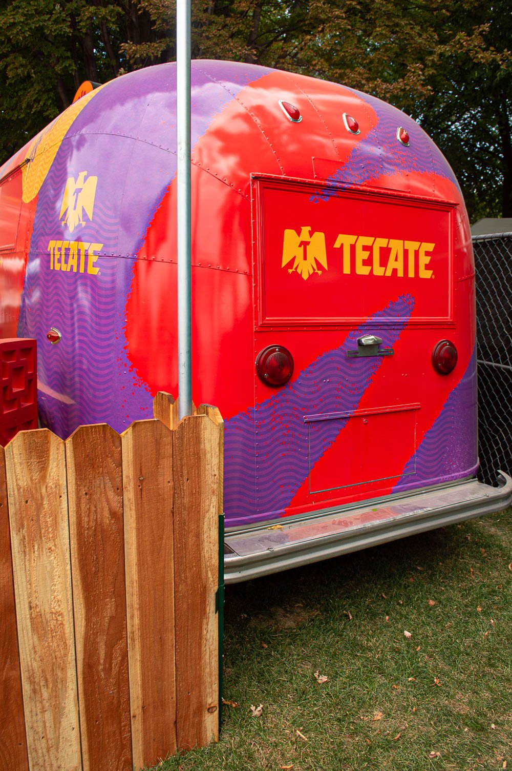 Caravana decorada con colores rojo, morado y amarillo, con el logo de Tecate en la parte delantera y lateral, ubicada en un área con árboles y cerca de una cerca de madera.