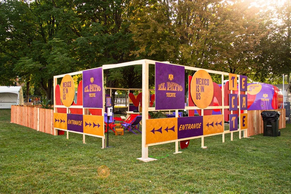 Estructura de madera decorada con cartel de colores que dice "México is in us", "El Patoite" y "Tecate" en un parque con árboles y césped.