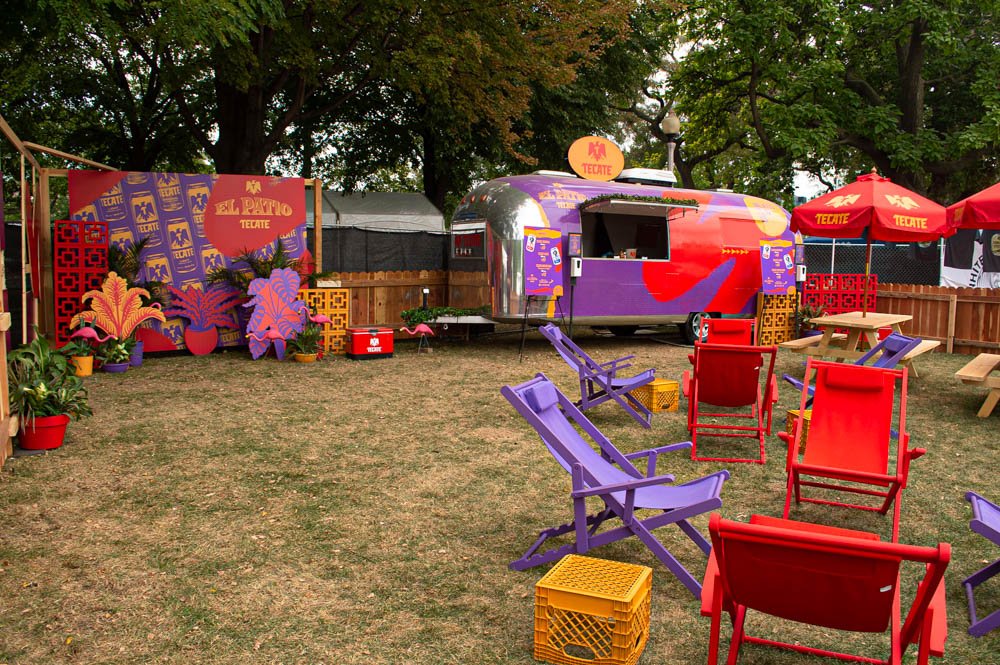 Área de sombra con sillas de colores, estructura de comida móvil de colores vibrantes, decoración con plantas y un cartel que dice 'El Patio Tecate', en un entorno con árboles y una cerca de madera.
