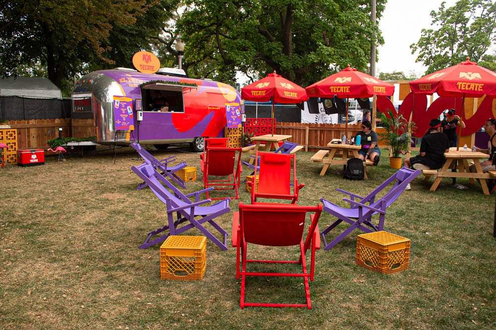 Área de fiesta al aire libre con mesas, sillas y sombrillas rojas con logo de Tecate, una camioneta decorada con colores vivos, y un grupo de personas sentadas en las mesas.
