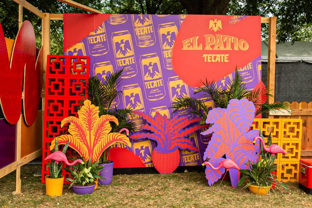 Decoración colorida con carteles y plantas tropicales, incluyendo flamingos de plástico, en un evento al aire libre denominado 'El Patio Tecate'.