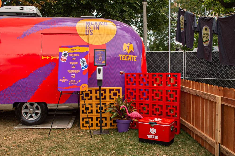 Carpa con decoración de Tecate, cartel de mensajería, florero con planta, flamingo decorativo, y ropa colgada en la cuerda en un espacio al aire libre