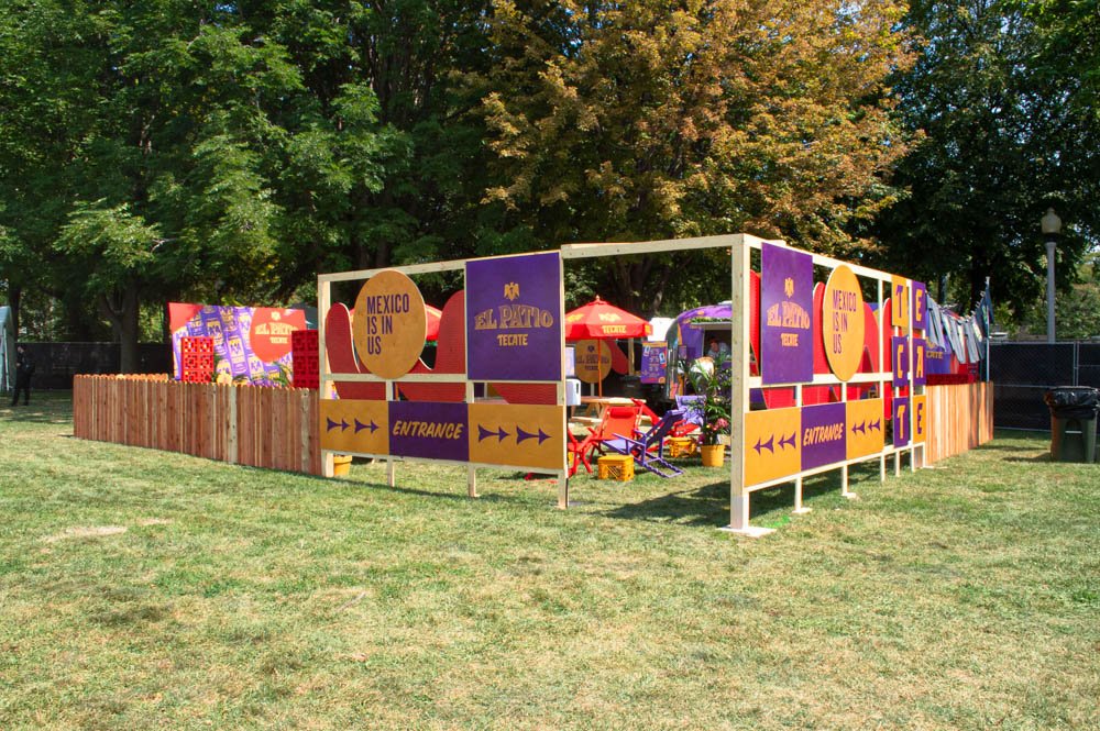 Entrada decorativa para una feria con colores vibrantes y carteles que dicen "El Patio," "México está en nosotros," y "Entrada" en un parque con árboles verdes.