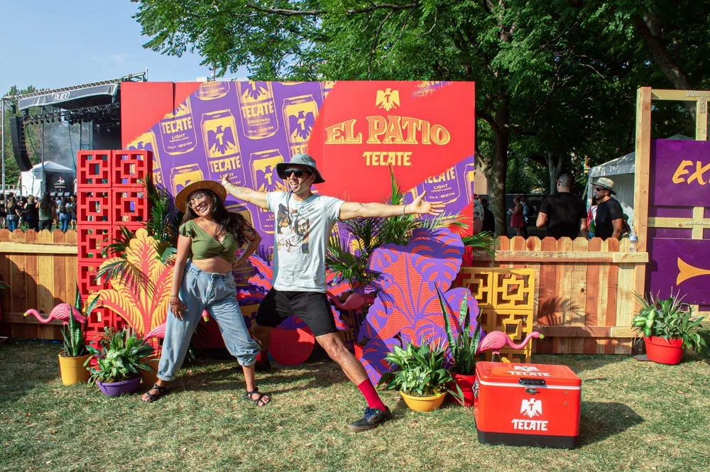 Dos personas posando en un evento de celebración con decoración colorida de flores, plantas y un cartel gigante que dice "El Patio Tecate" en un entorno de festival al aire libre.