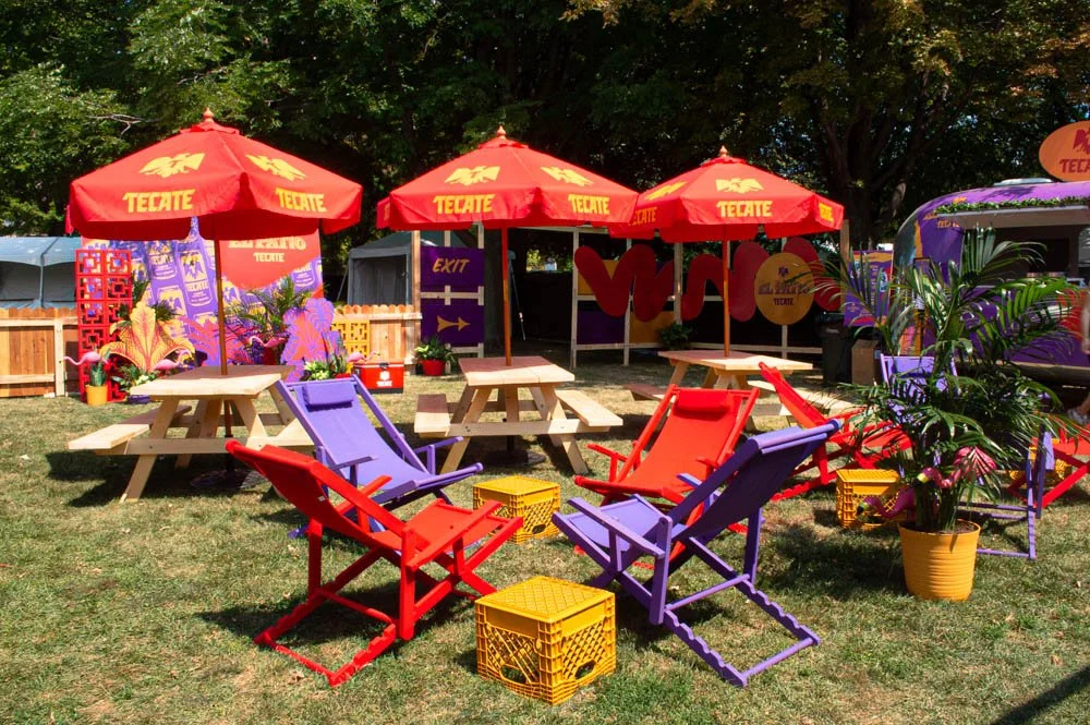 Área de descanso con mesas de picnic, sillas y sombrillas rojas en un parque, rodeado de árboles y decoraciones coloridas.