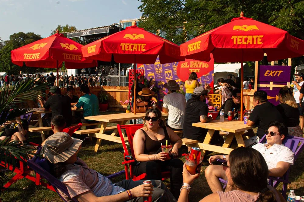 Personas en un festival al aire libre sentado bajo sombrillas rojas con el logo de Tecate, disfrutando de bebidas y música en un ambiente festivo.