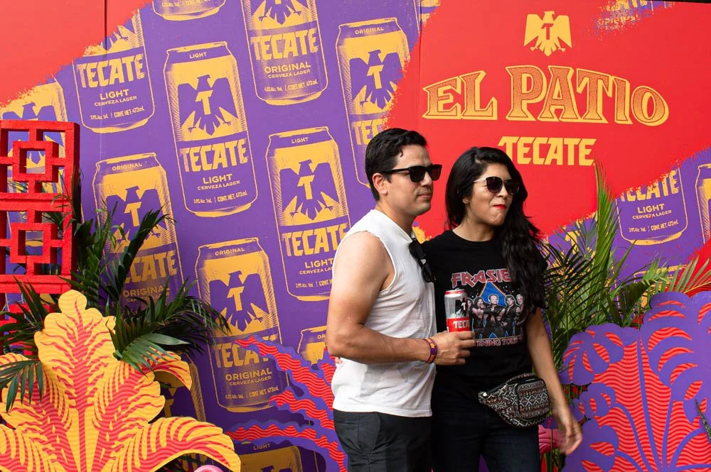 Dos personas posando frente a un fondo con logos de Tecate y decoración colorida con plantas.