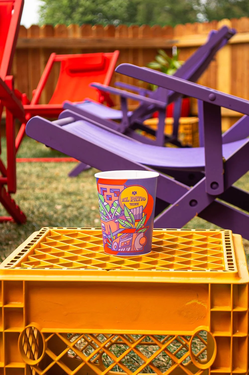 Vasos y sillas de colores en un jardín, un vaso con el logo de 'El Patio TECATE' en primer plano sobre un contenedor naranja.