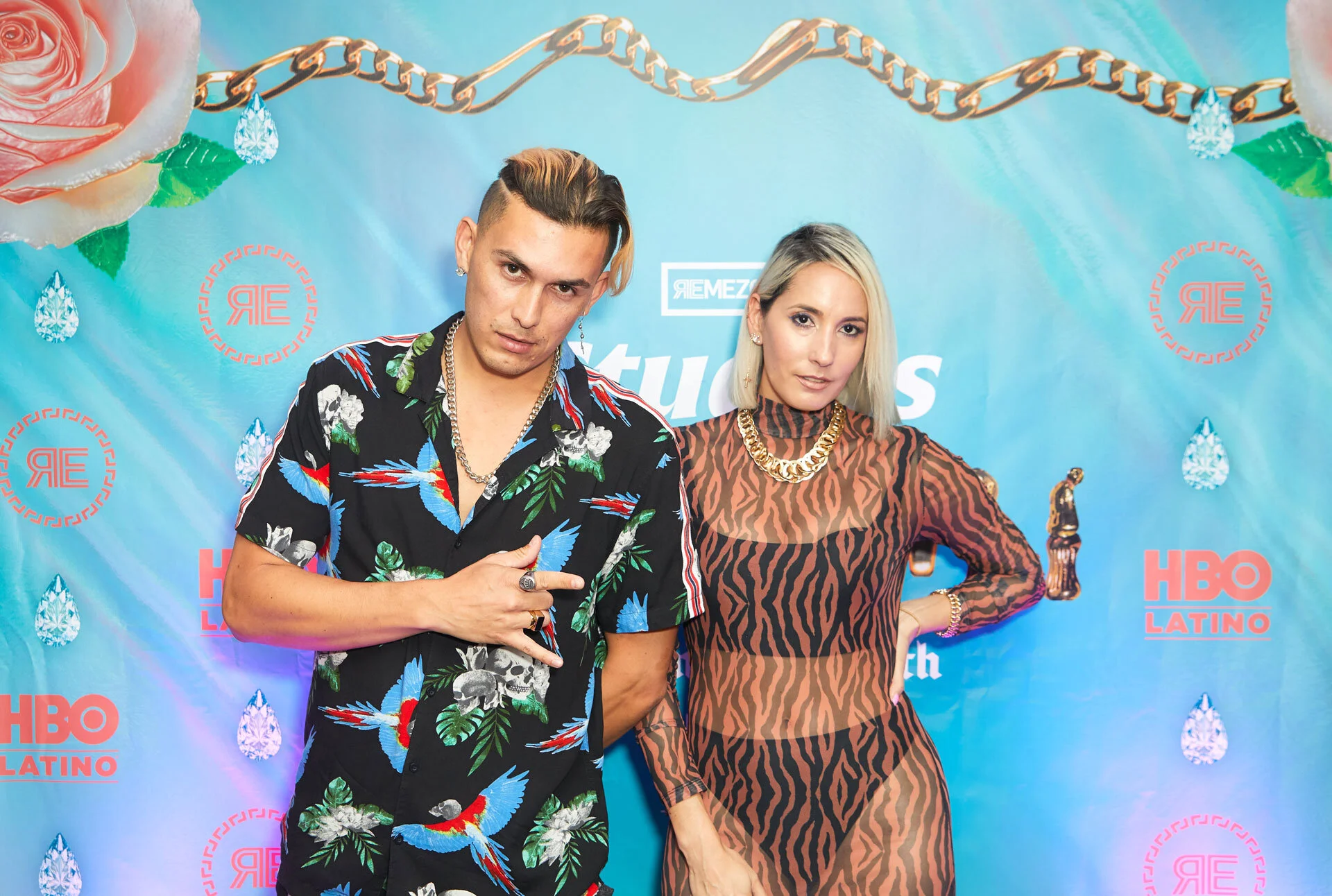Dos personas posando en un evento con fondo azul, decoraciones de diamantes y rosas, y logotipos de HBO Latino y otros logos.