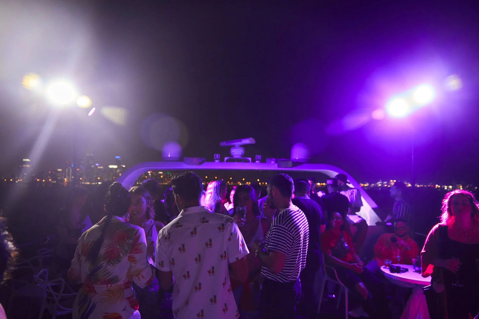 Grupo de personas en una fiesta en la noche, con luces moradas y fondo de ciudad