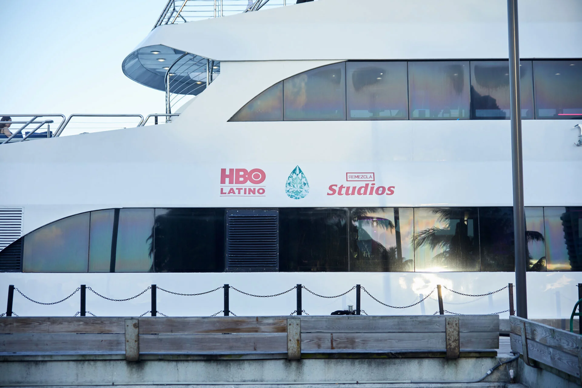 Un barco grande y moderno con logotipos de HBO Latino y Estudios está atracado en el puerto, con una cerca y un muro de madera en primer plano, y palmeras reflejadas en las ventanas.