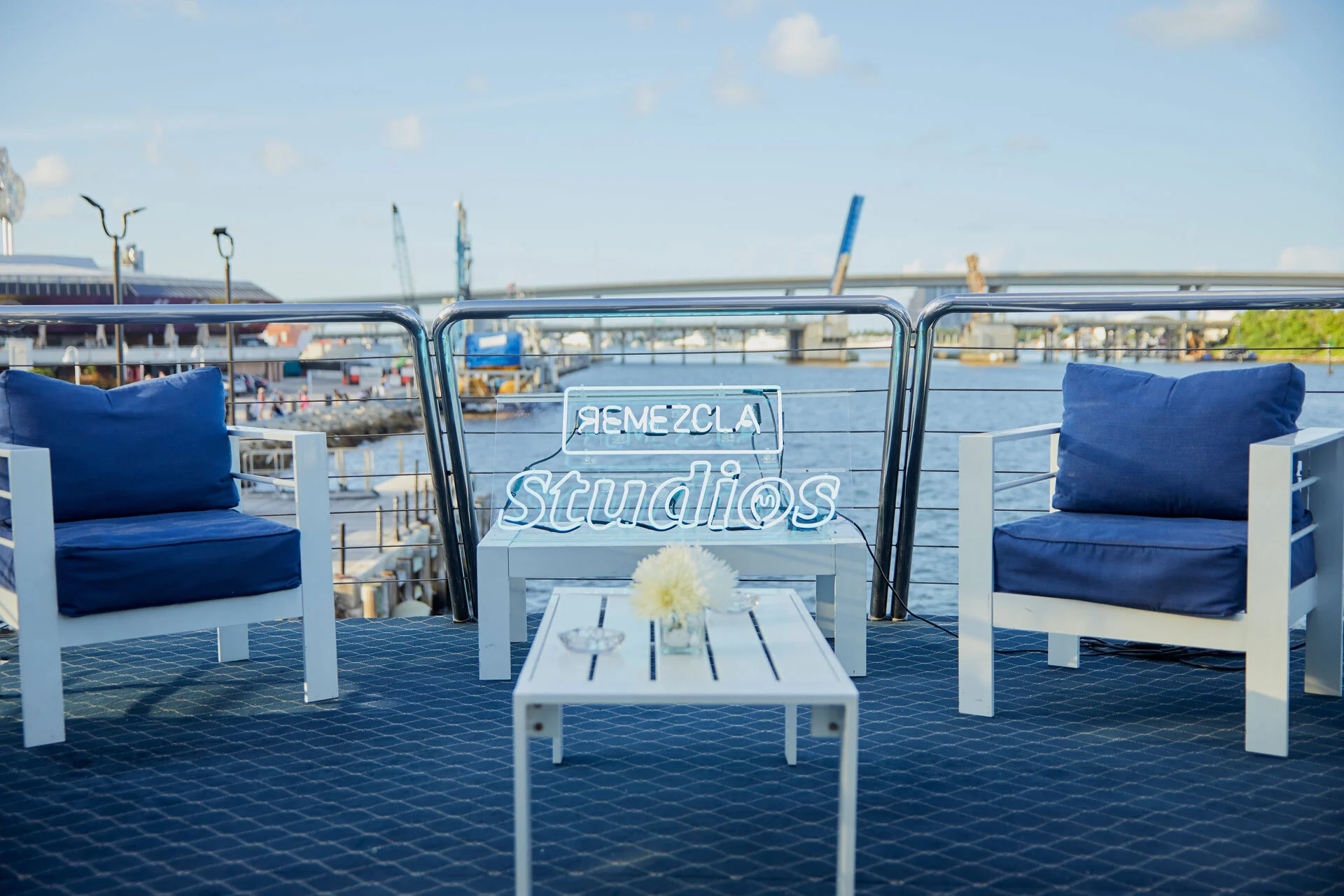 Área de descanso en un muelle con sillas blancas y cojines azules, una mesa de blanco con una flor y una señal de neón que dice 'Remezcla Studios' frente a un río, con edificios y barcos en el fondo bajo cielo soleado.